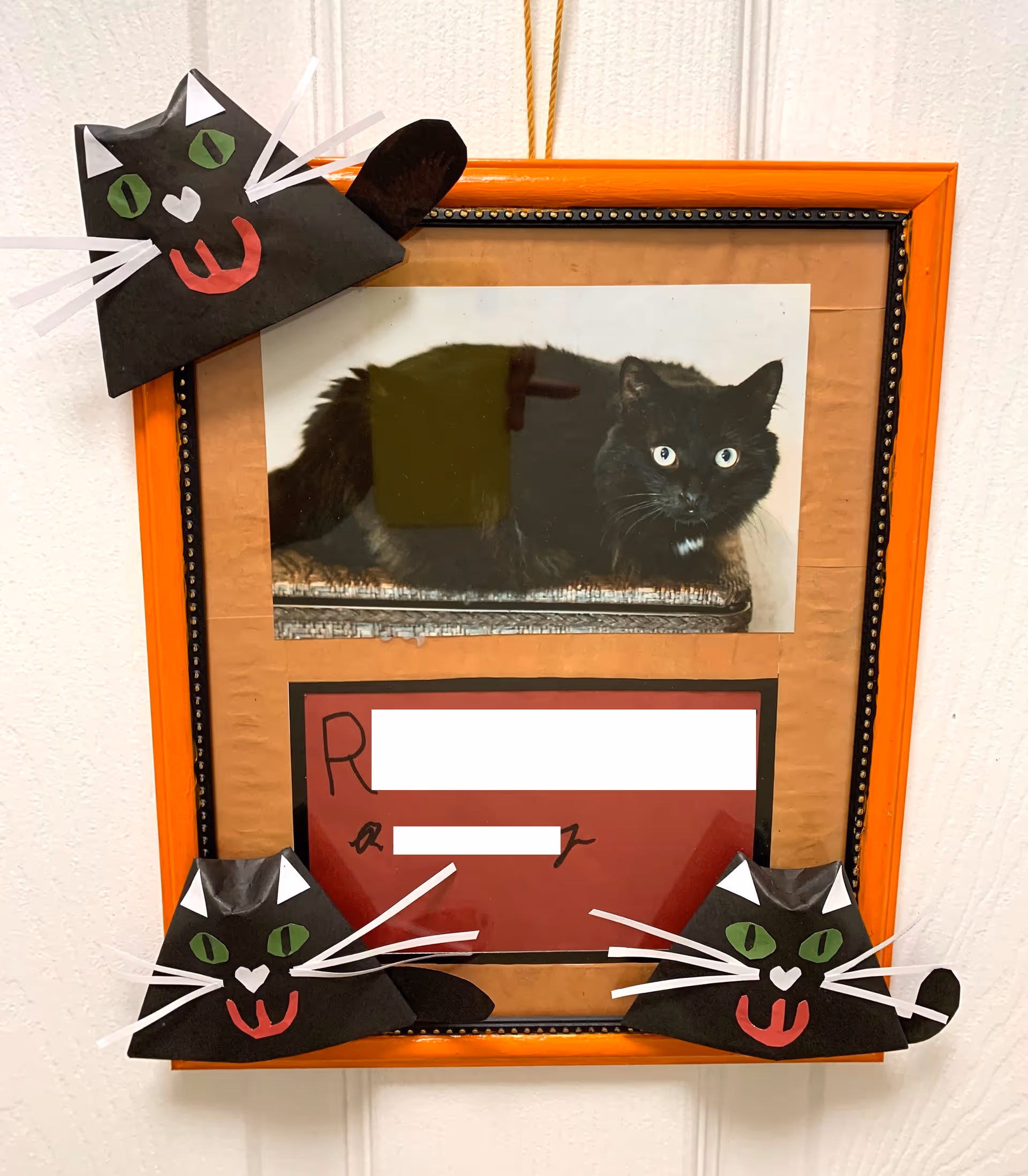 A framed photo of a black cat with green eyes lying down, decorated with three black paper cat faces with green eyes, white whiskers, and red tongues attached to the top left, bottom left, and bottom right corners of the frame. The frame is orange with a black inner border and is hanging on a white wall.