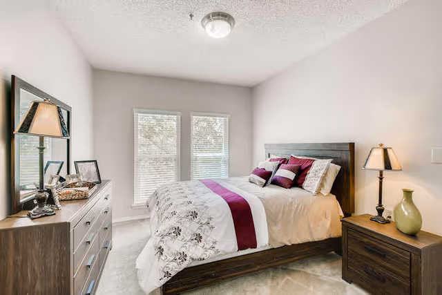 A bright bedroom with a large bed featuring white and purple bedding, two windows with blinds letting in natural light, a wooden dresser with a mirror and decorative items, and a wooden nightstand with a lamp and a green vase.