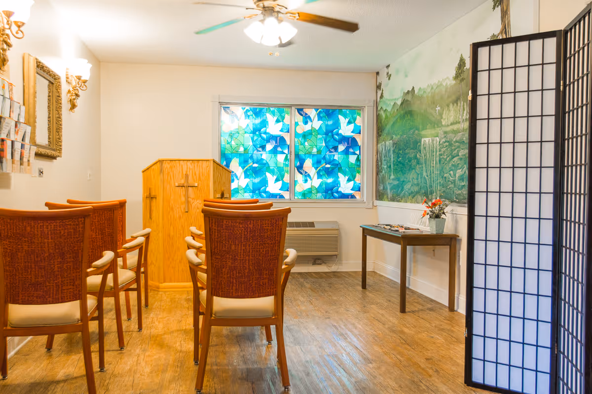 Small chapel-style room with a wooden pulpit, arranged chairs facing a colorful stained-glass window and a mural.