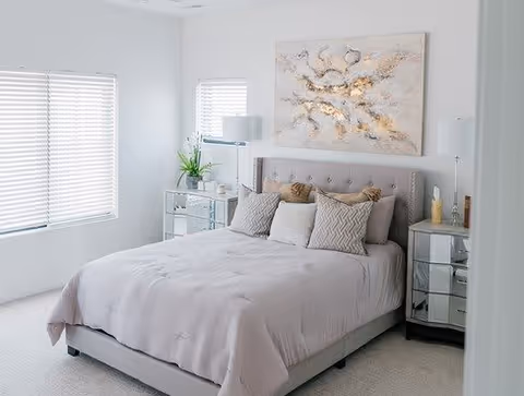A bright bedroom with a neatly made bed featuring light gray bedding and multiple pillows. The room has two mirrored nightstands on either side of the bed, each with a lamp and decorative items. There are two windows with white blinds allowing natural light to fill the space. A large abstract painting hangs above the bed.