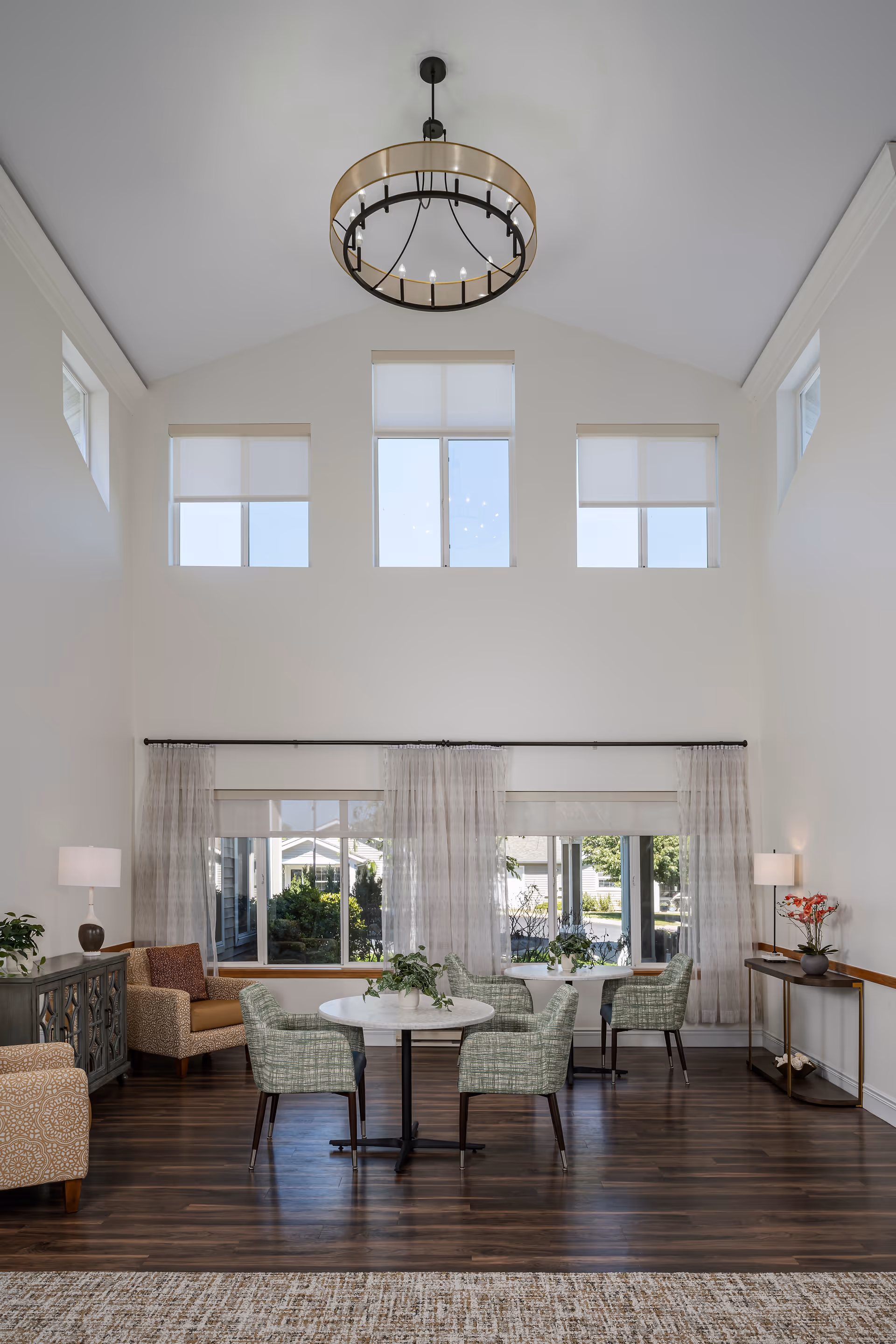 Bright, airy senior living common room with high ceilings, a chandelier, round tables and upholstered chairs in front of large windows.