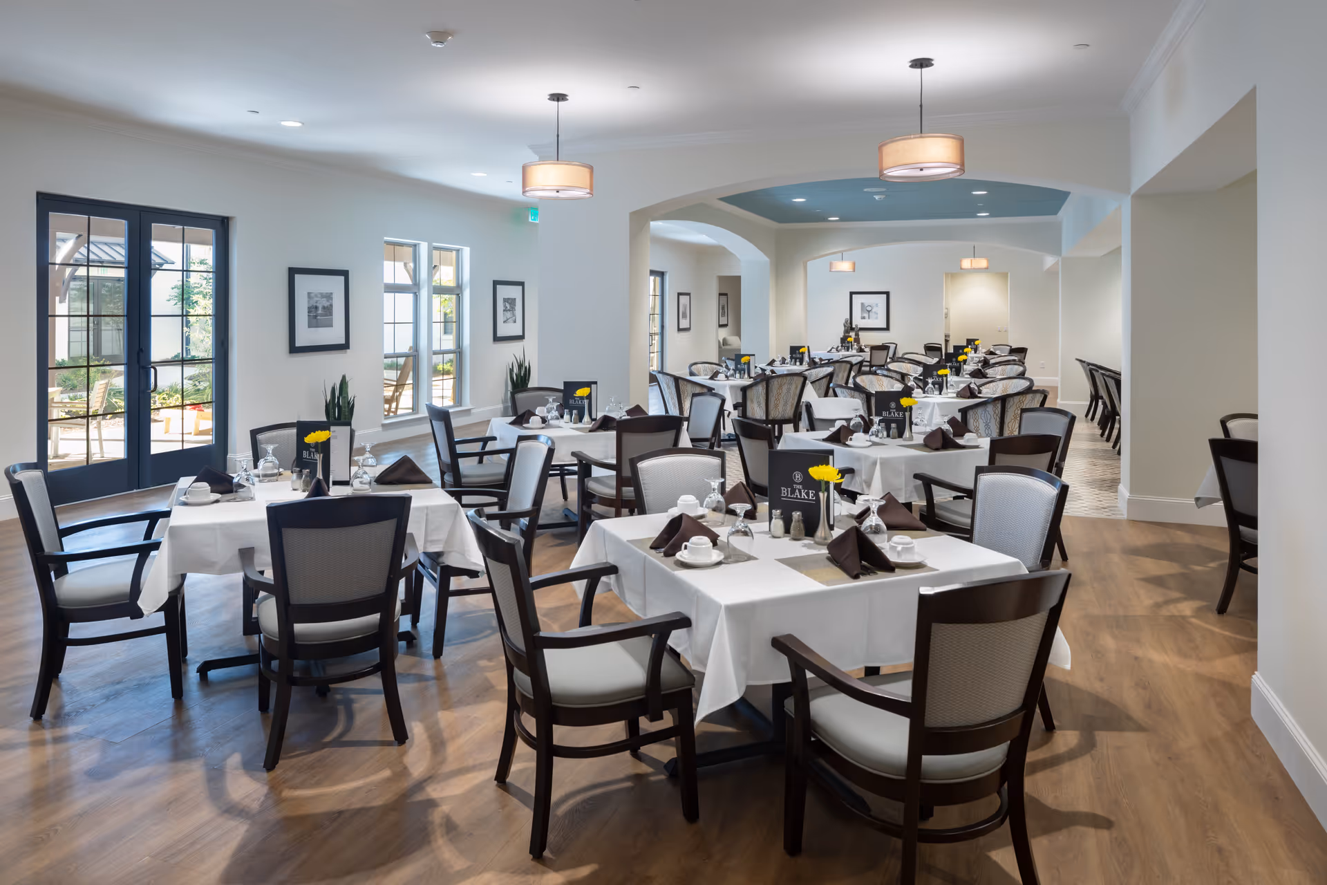 A bright and spacious dining room with multiple tables covered in white tablecloths, each set with cups, saucers, napkins, and small flower vases. The room features wooden chairs with cushioned seats, large windows, and glass doors allowing natural light to enter. The walls are decorated with framed black and white photos, and modern pendant lights hang from the ceiling.