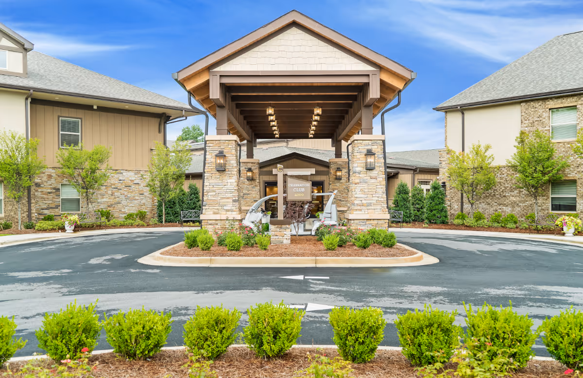 Front entrance of Celebration Village Forsyth featuring a covered driveway with stone pillars, landscaped bushes and flowers, and two buildings on either side under a blue sky.