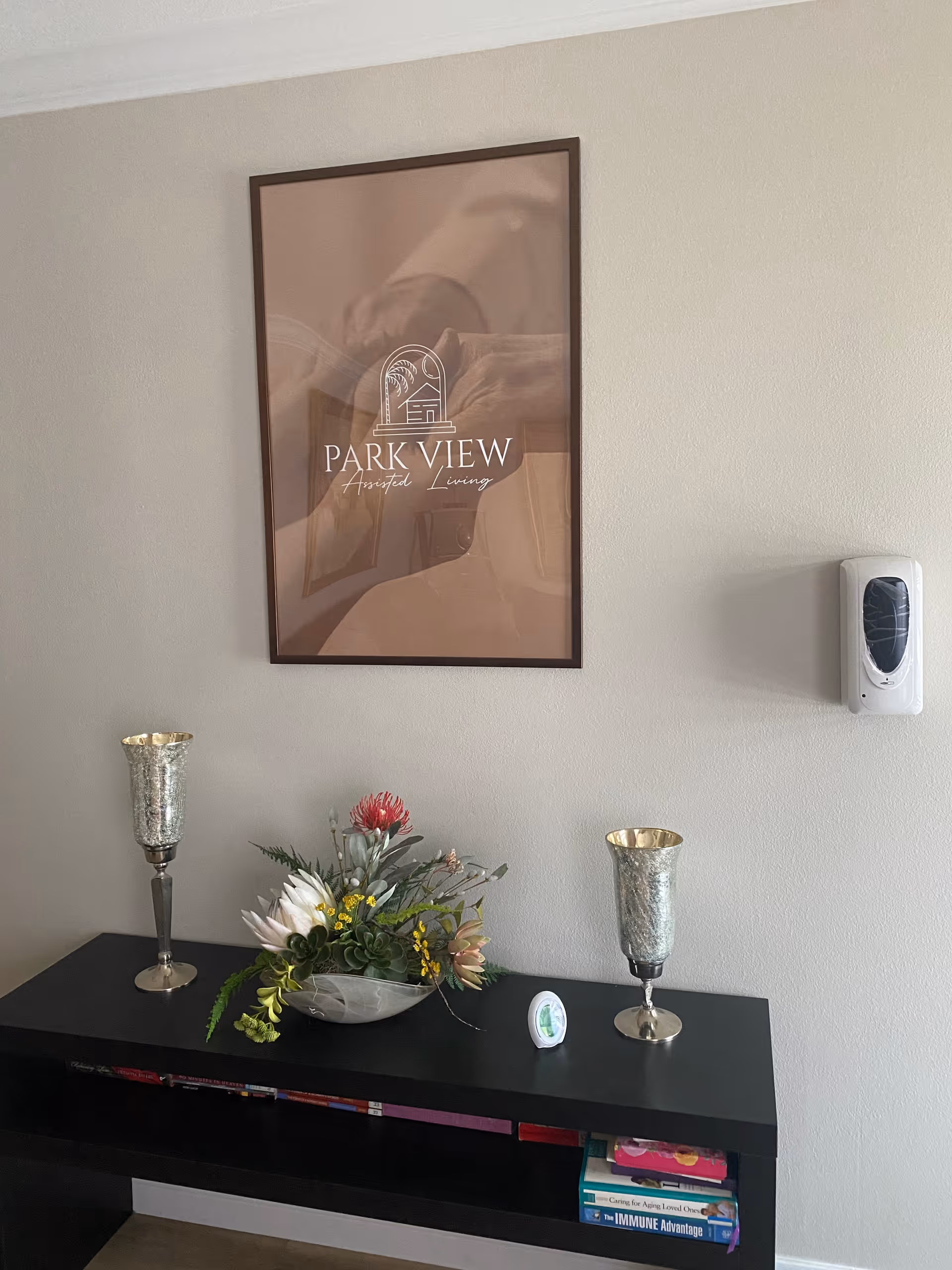 A beige wall with a framed poster that reads 'Park View Assisted Living' above a black console table. On the table are two silver goblets, a decorative floral arrangement, and a small round device. A wall-mounted hand sanitizer dispenser is visible to the right of the poster.
