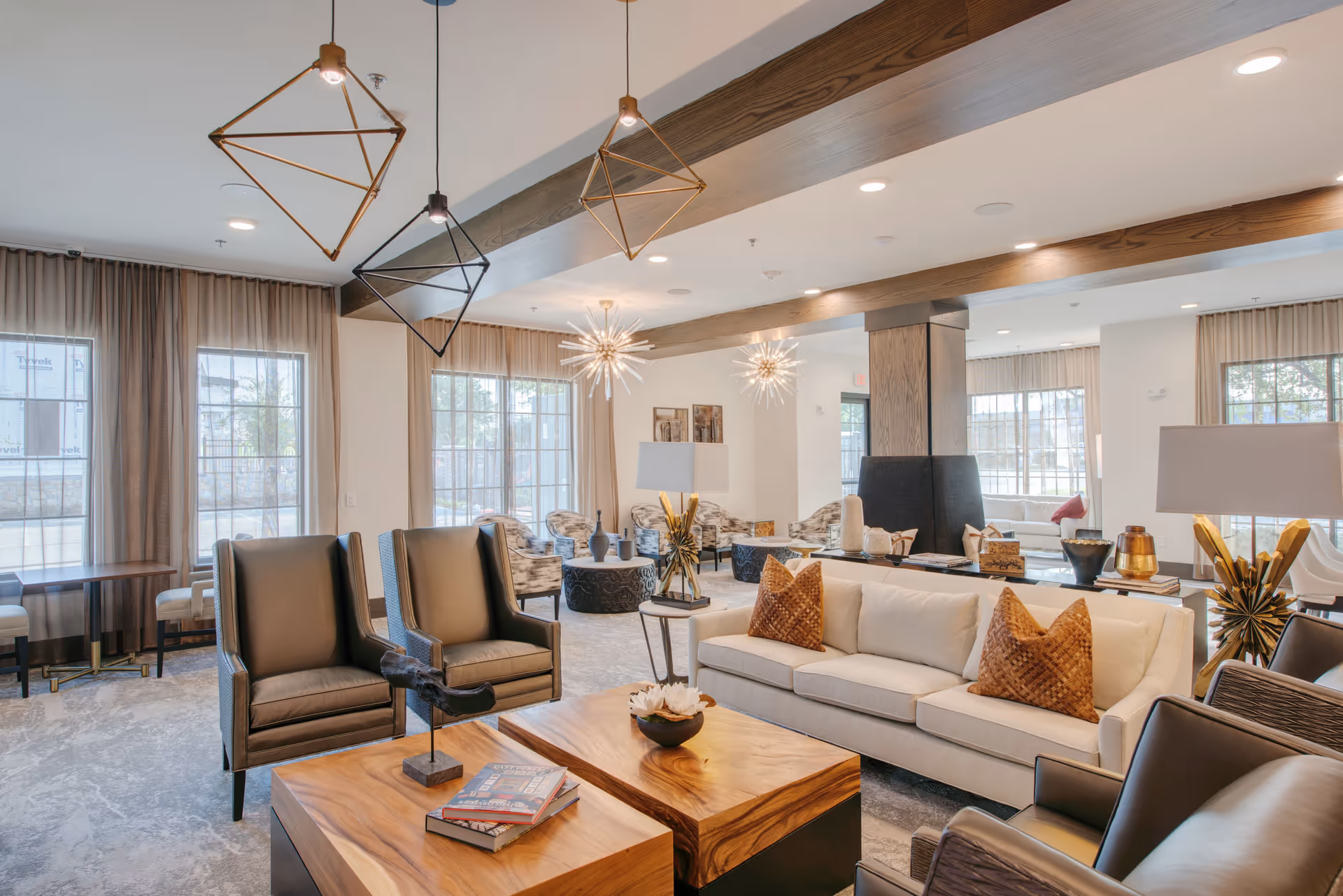 A spacious and modern living room area in Everleigh Forestwood 55+ Apartment Homes featuring a beige sofa with brown pillows, two leather armchairs, wooden coffee tables with decorative items, large windows with curtains, and unique geometric pendant lights hanging from the ceiling.