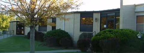 Single-story brick facility with large windows, trimmed shrubs, and a tree in front.