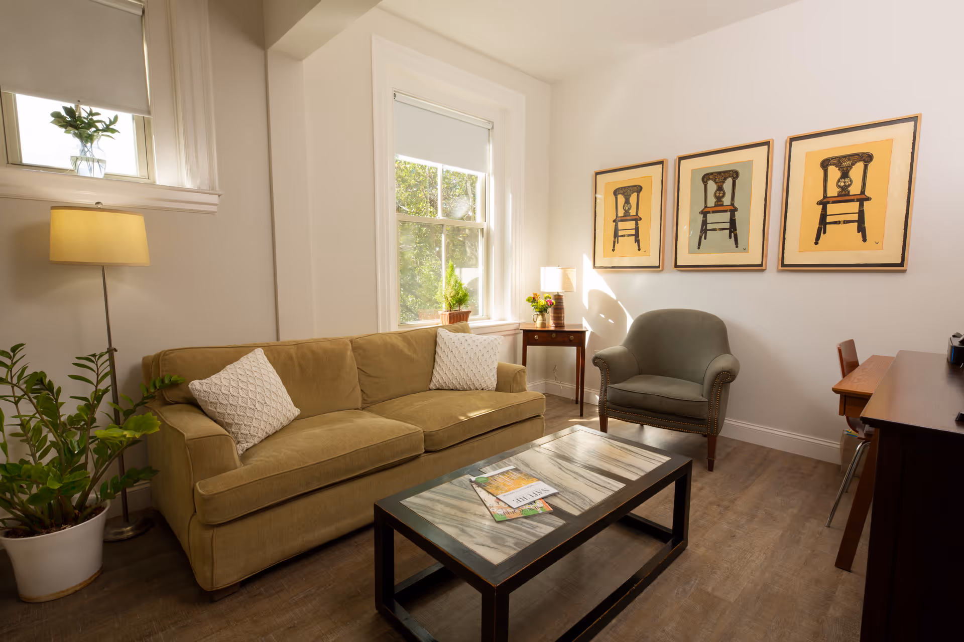 A cozy living room with a beige sofa adorned with two white pillows, a green armchair, and a rectangular coffee table with magazines on it. The room has wooden flooring, a floor lamp, a small side table with a lamp and flowers, and three framed pictures of chairs on the wall. There are two windows letting in natural light, and a potted plant is placed near the sofa.