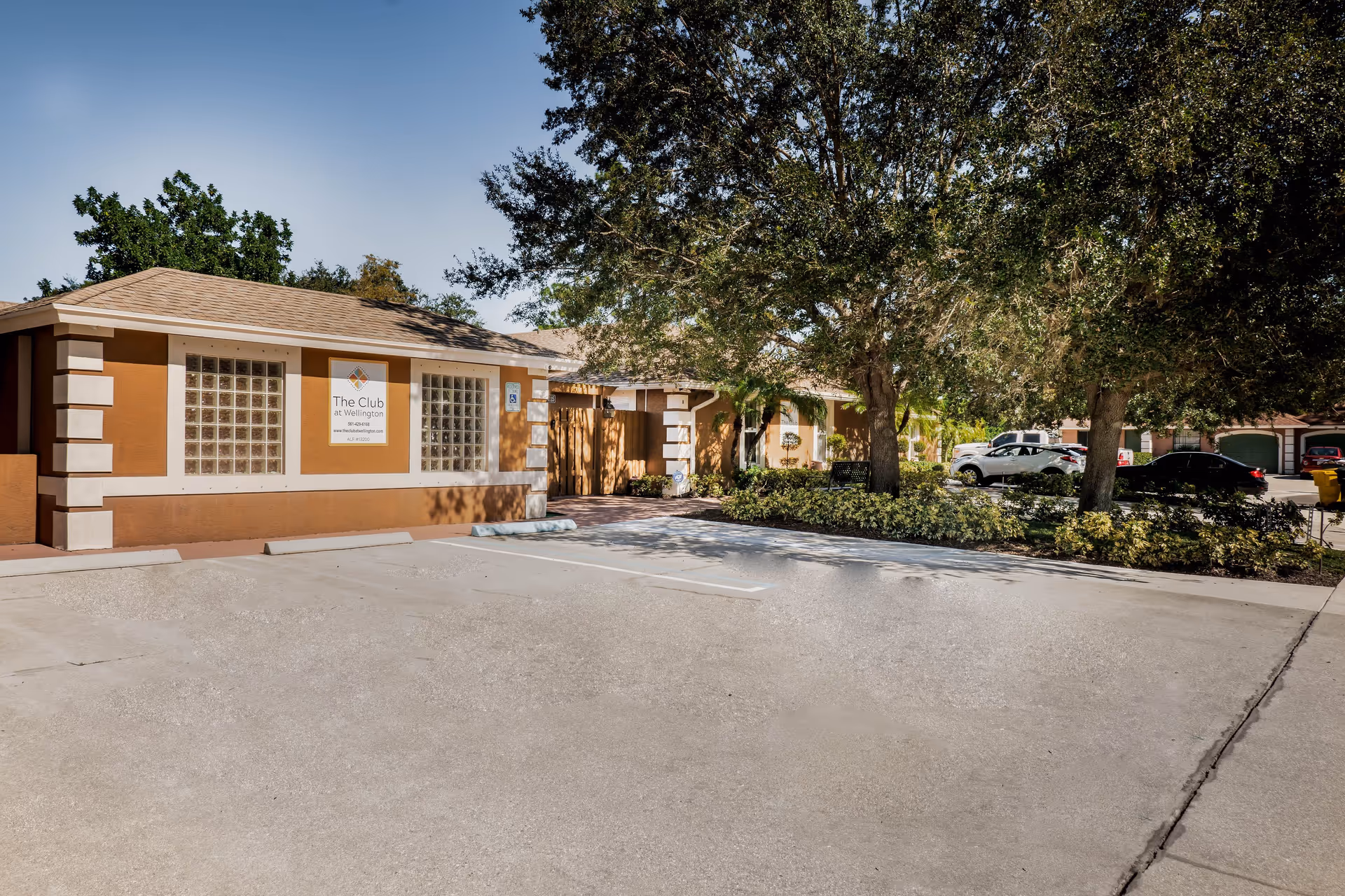 Exterior view of The Club at Wellington assisted living and memory care facility showing a single-story building with a brown and beige facade, glass block windows, a sign with the facility name, a parking area with marked spaces, trees, and shrubs.