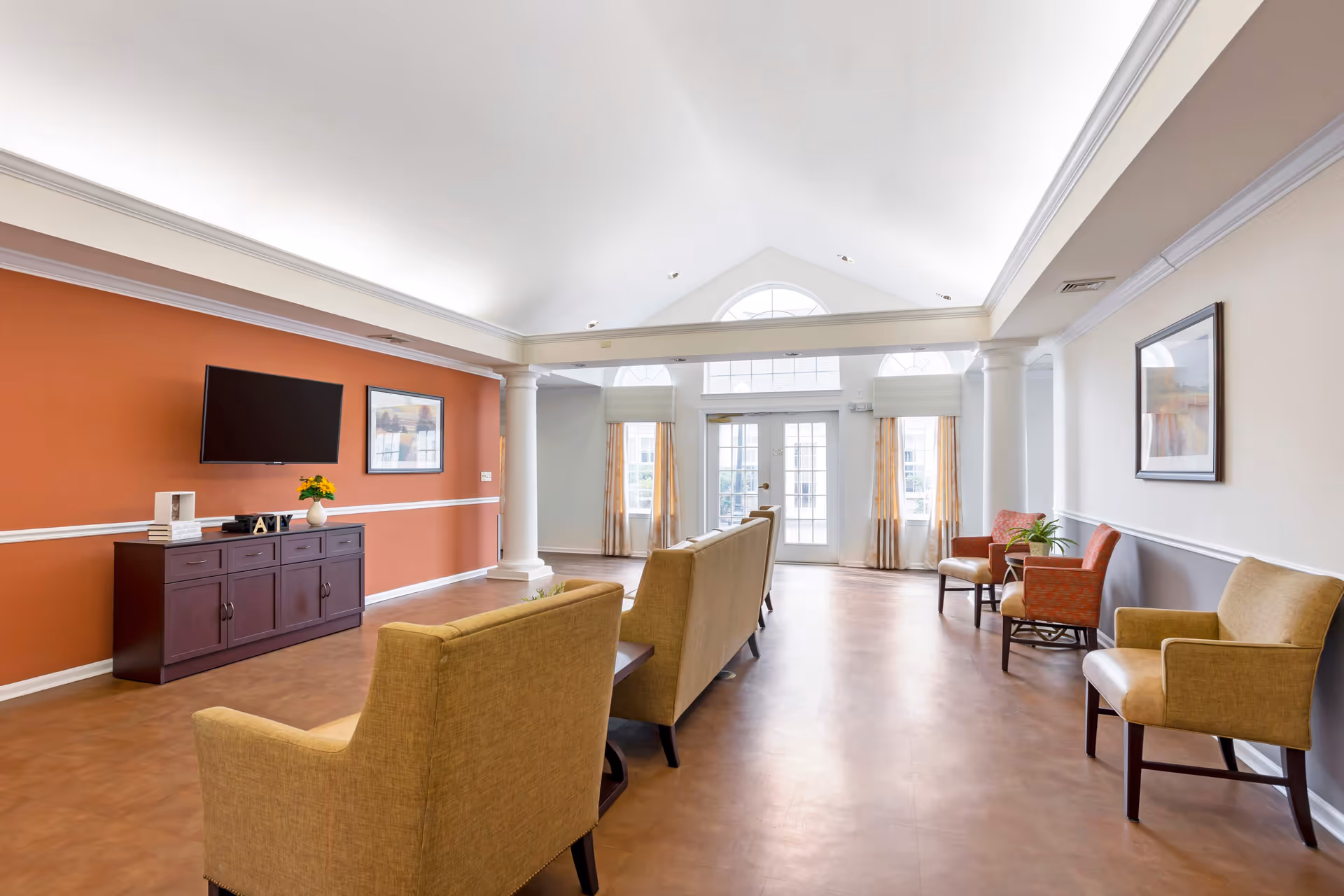 A bright and spacious living room area in a senior living facility with beige armchairs arranged in rows facing a wall-mounted flat screen TV. The room features a high vaulted ceiling, large windows with curtains, white columns, and framed artwork on the walls. There is a dark wooden cabinet beneath the TV with decorative items on top.