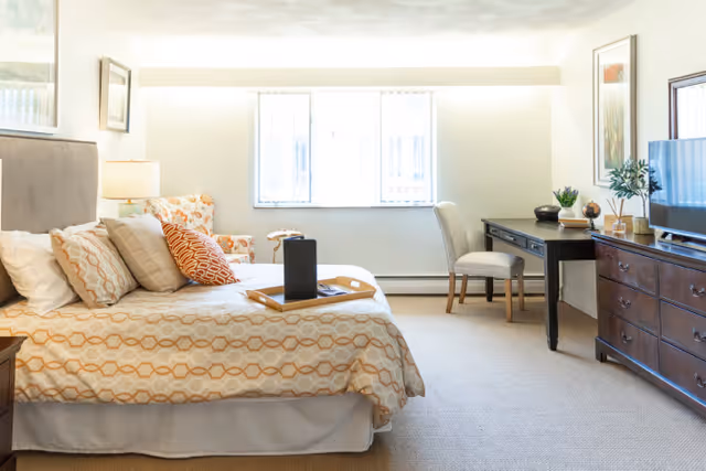 A bright and cozy bedroom in a senior living facility featuring a bed with patterned bedding and multiple pillows, a bedside lamp, an armchair with a floral pattern, a wooden desk with a chair, a flat-screen TV on a wooden dresser, and framed artwork on the walls. Two windows allow natural light to fill the room.