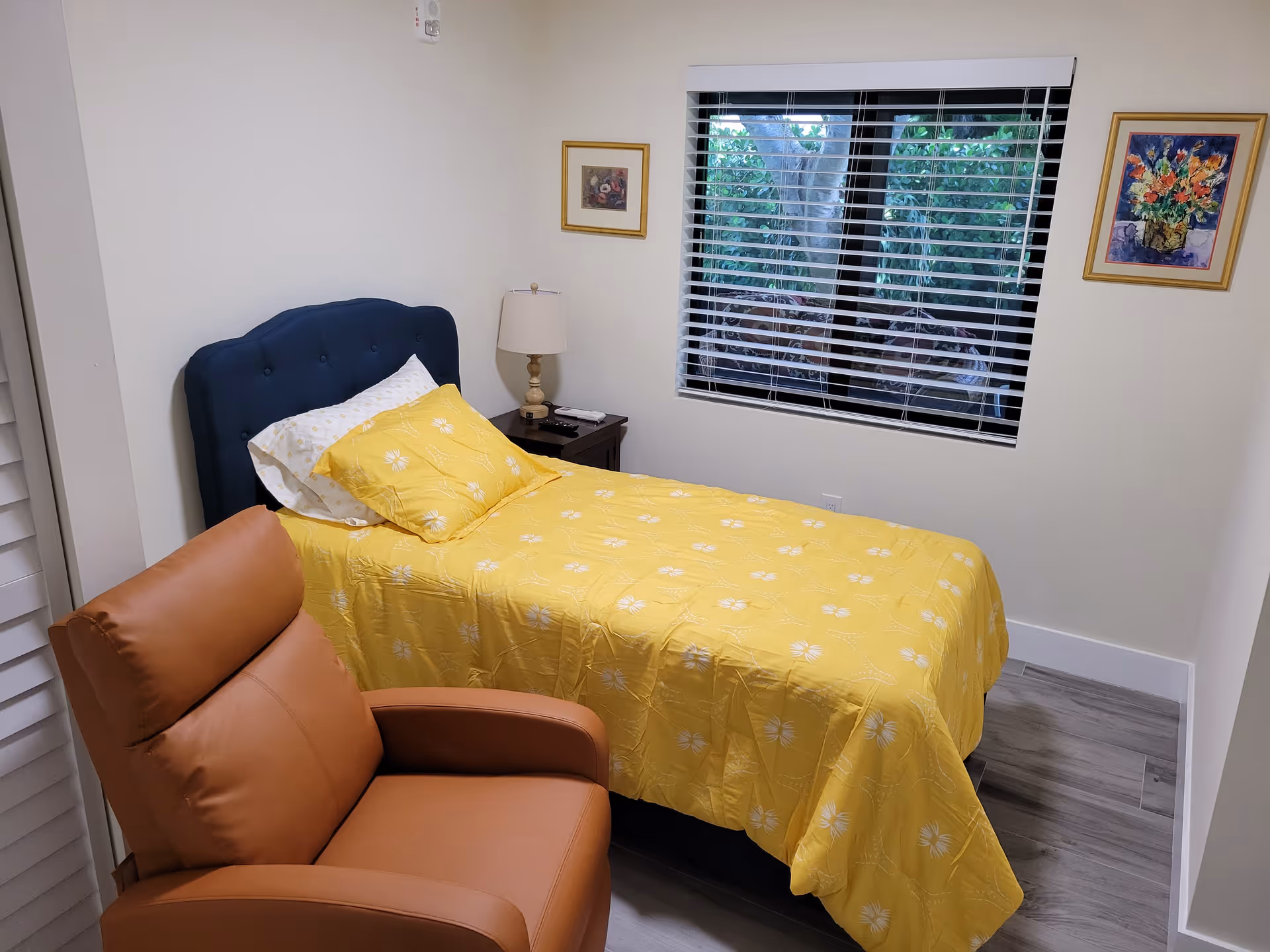A small bedroom with a single bed covered in a yellow bedspread with white floral patterns. The bed has a dark blue tufted headboard and white pillows. Next to the bed is a dark wooden nightstand with a beige lamp and some items on it. A brown leather recliner chair is positioned in the foreground. The room has light-colored walls, two framed floral paintings, and a window with white blinds showing greenery outside.