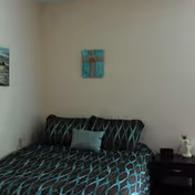 A bedroom with a bed covered in a dark blue and black patterned comforter and matching pillows. There is a small light blue pillow in the center. On the wall above the bed hangs a decorative piece featuring a cross. To the right of the bed is a dark nightstand with a small lamp and a few items on it. The walls are plain and light-colored.