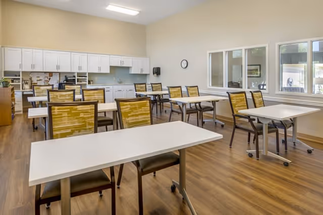 A bright room with multiple tables and chairs arranged in rows on a wooden floor. The chairs have yellow patterned backs and dark frames. The back wall features white cabinets and a countertop with a sink. There are windows on the right wall letting in natural light, and a clock is mounted on the wall above the countertop.