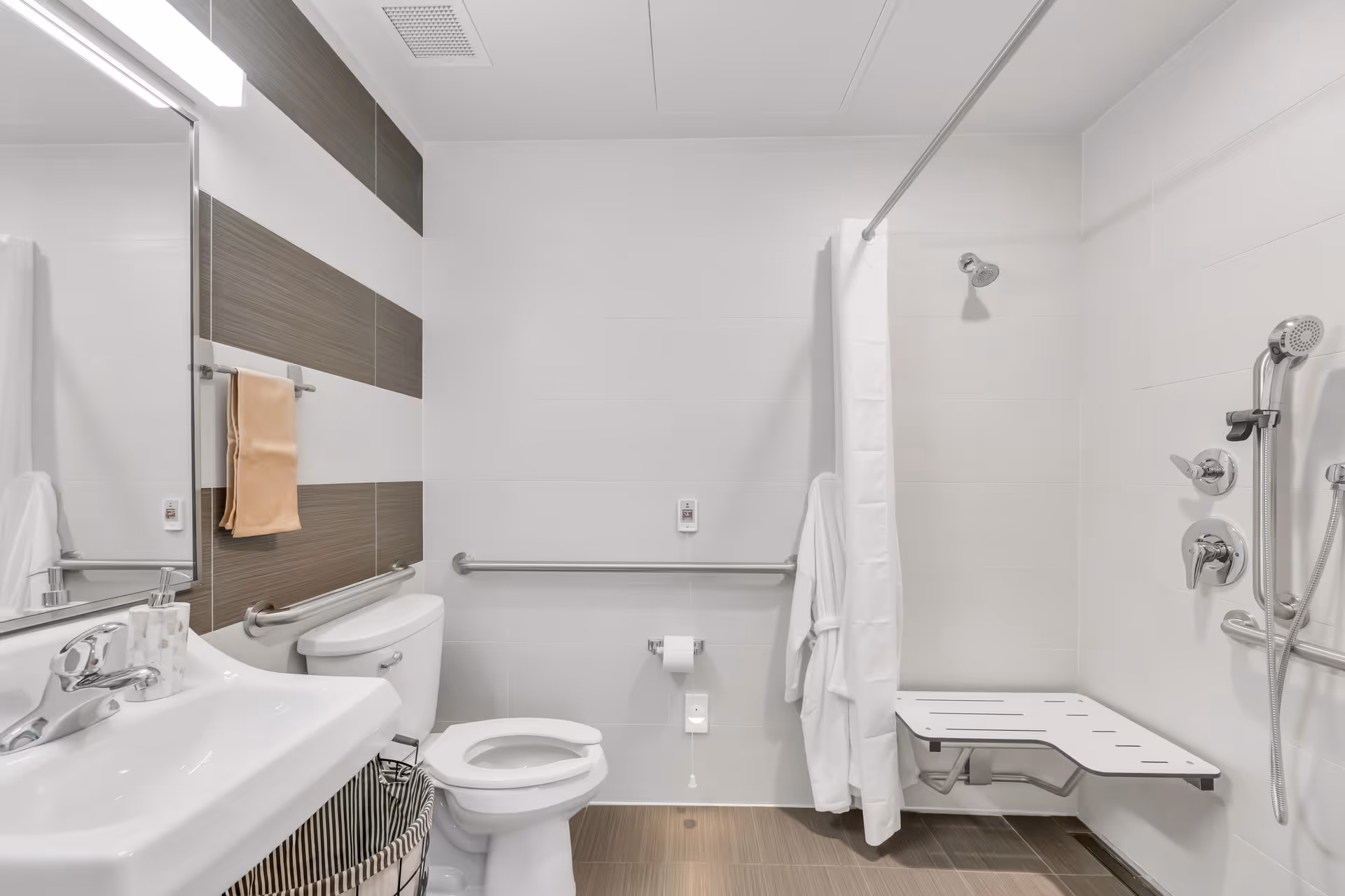 Accessible bathroom with a sink, toilet, grab bars, shower curtain, wall-mounted fold-down shower bench, and handheld showerhead.