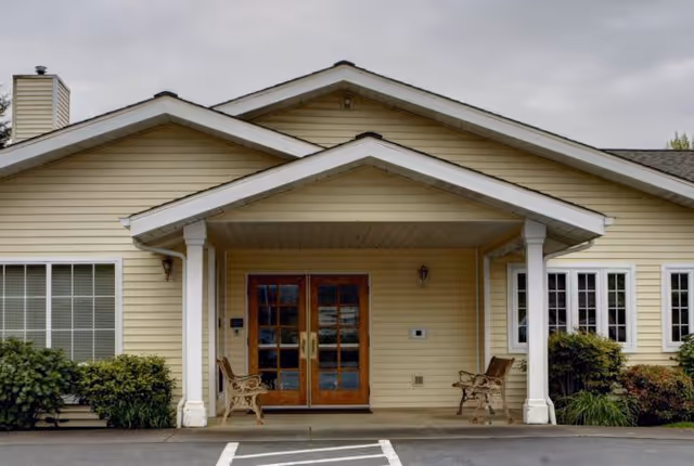 Front exterior view of a single-story senior living facility building with beige siding, a covered entrance supported by white columns, double wooden doors with glass panels, two benches on either side of the entrance, and surrounding greenery.