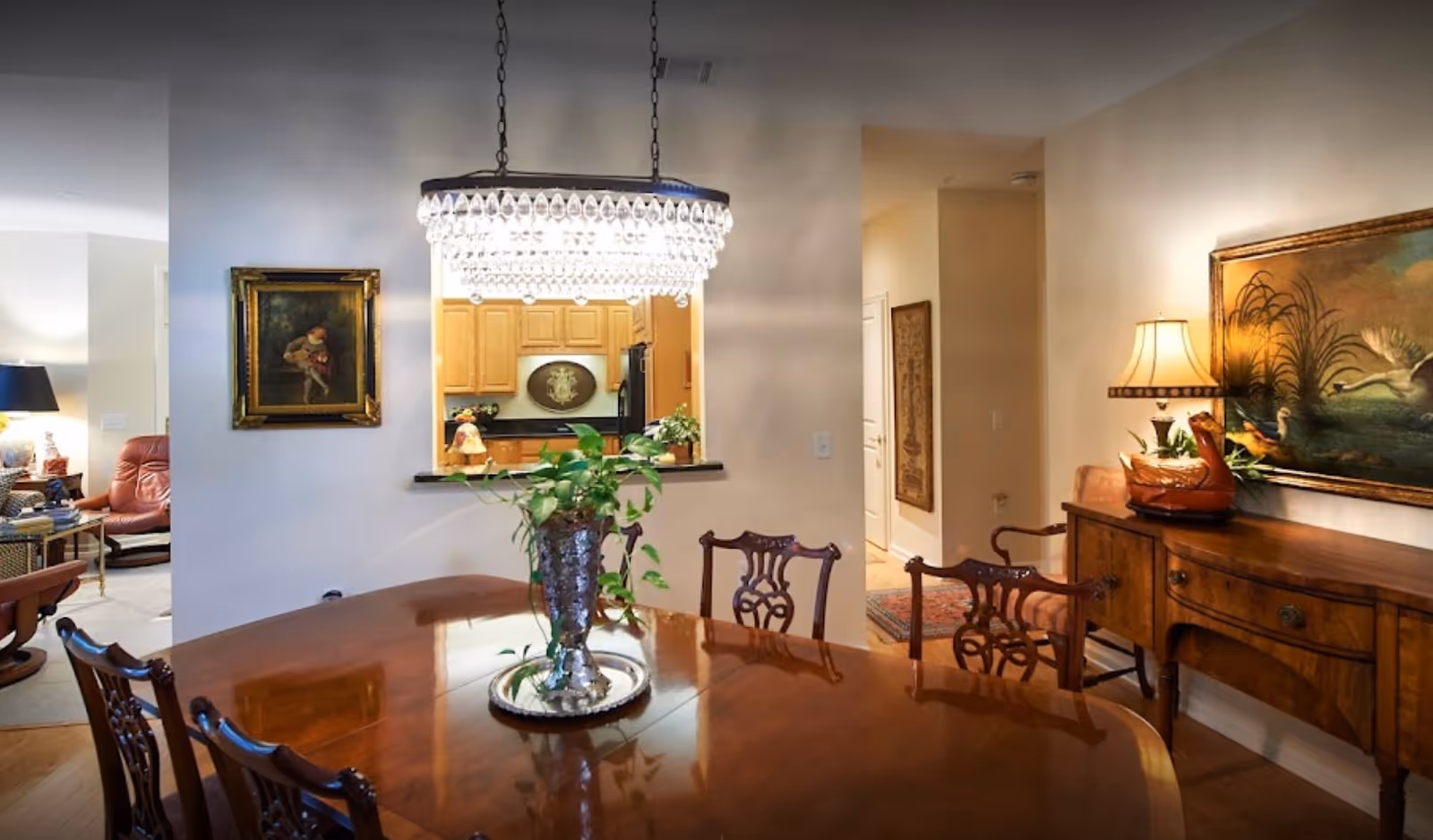 A dining room with a polished wooden table and ornate wooden chairs. A crystal chandelier hangs above the table. There is a silver vase with green plants on the table. In the background, a kitchen with wooden cabinets is visible through a pass-through window. The room is decorated with framed paintings on the walls and a wooden sideboard with a lamp and decorative items.