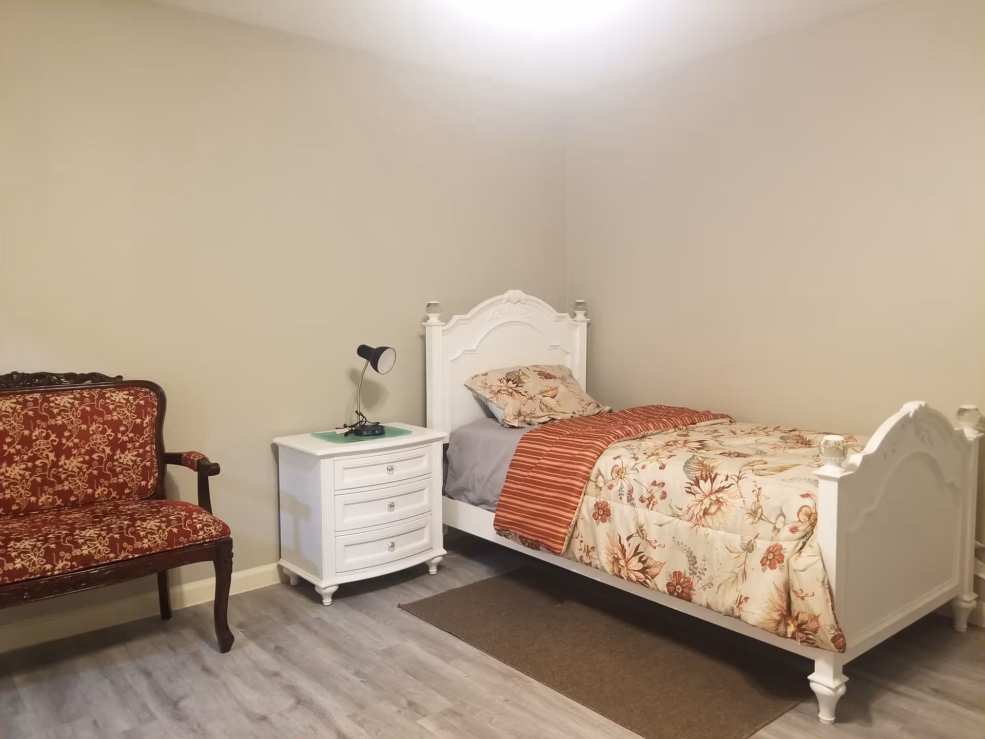 A white bed frame with an ornate headboard, floral bedding, nightstand, lamp, and a patterned chair.