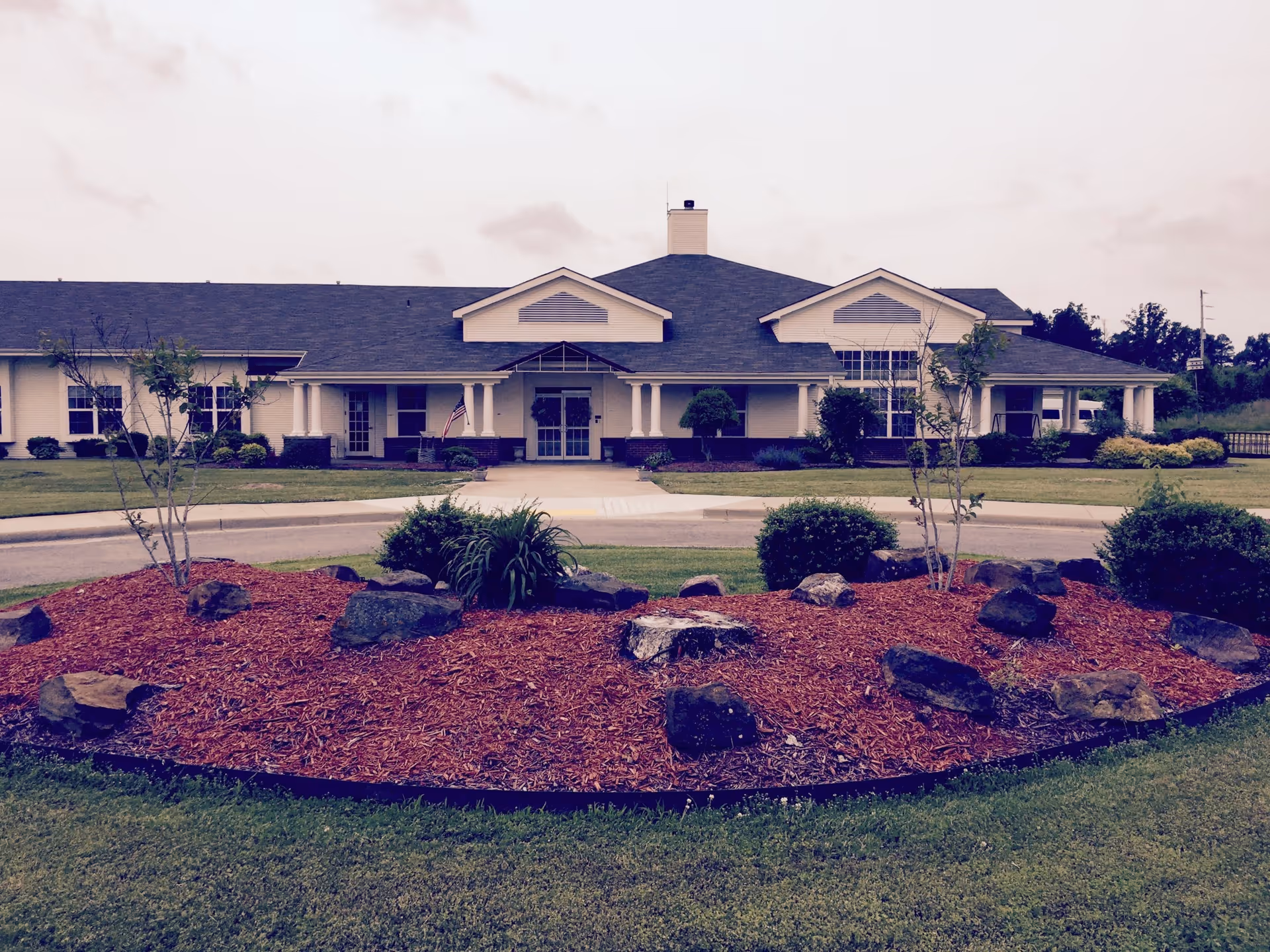 Front exterior view of StoneBridge Senior Living - Pocahontas building with a landscaped area featuring mulch, rocks, and small bushes in the foreground, and a circular driveway leading to the entrance.