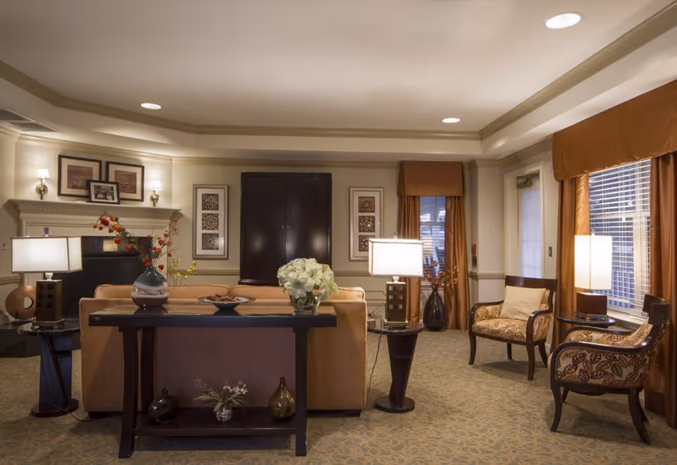 A cozy living room area in a senior living facility with a beige sofa, two patterned armchairs, and several lamps providing warm lighting. The room features a fireplace with framed pictures above it, a dark wooden console table behind the sofa with decorative vases and flowers, and large windows with brown curtains allowing natural light to enter.