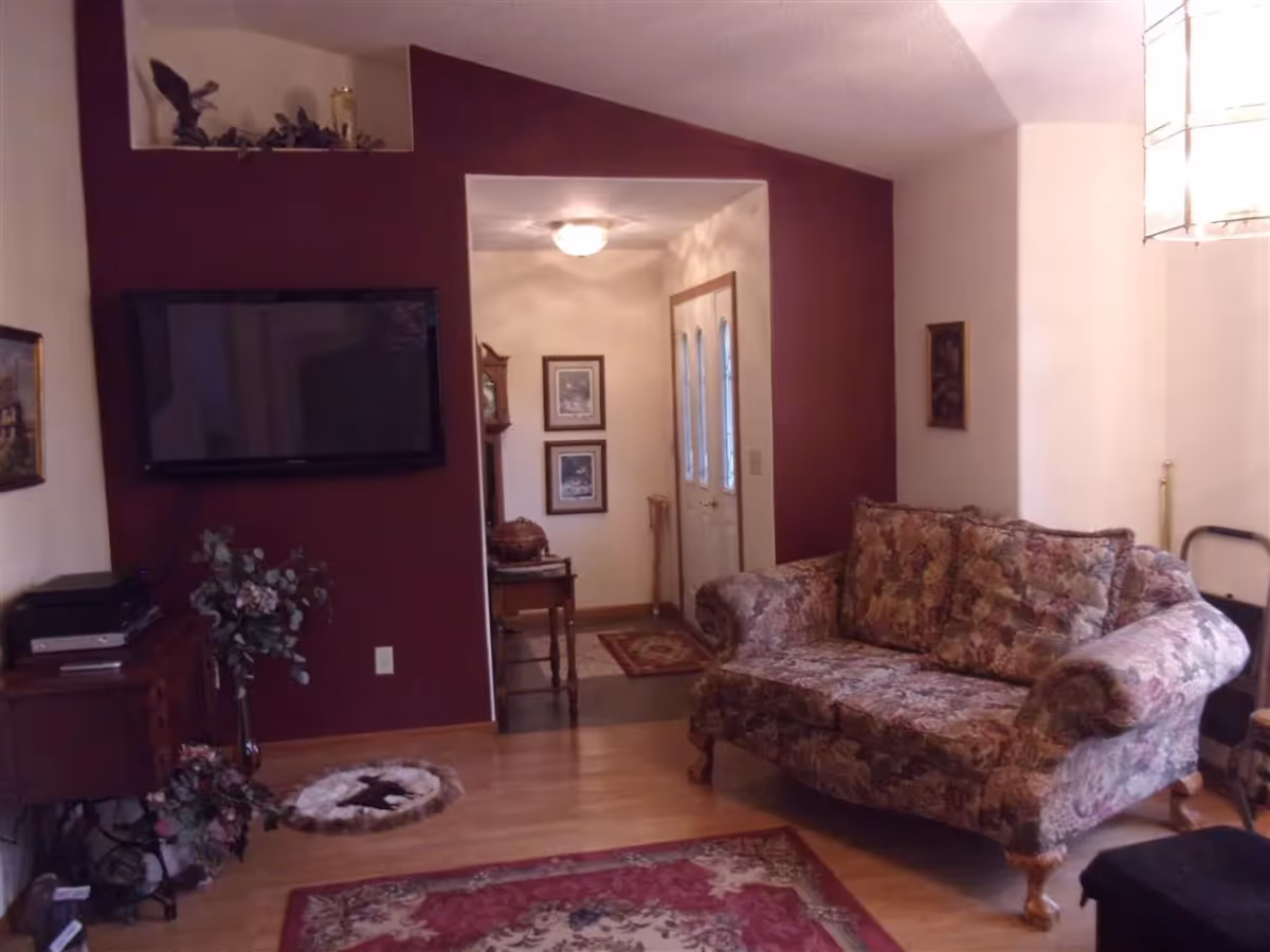 A cozy living room with a floral patterned sofa, a wall-mounted flat screen TV on a maroon accent wall, wooden flooring, and a small entryway with framed pictures and a grandfather clock.