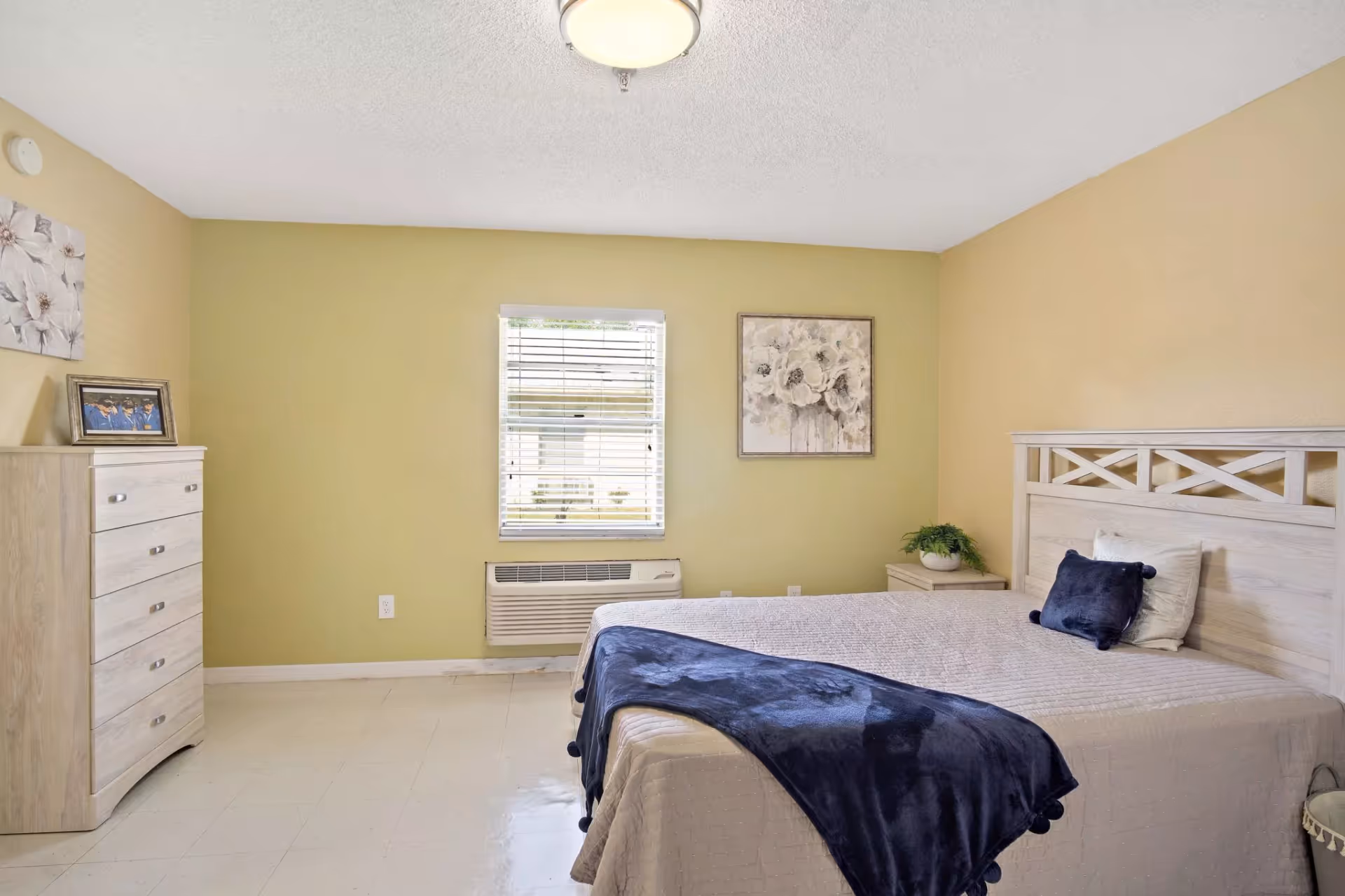 A bedroom with light yellow walls, a window with blinds, a bed with beige bedding and a dark blue throw blanket, a light wood headboard, a matching dresser with a framed photo on top, a nightstand with a potted plant, and floral artwork on the walls.
