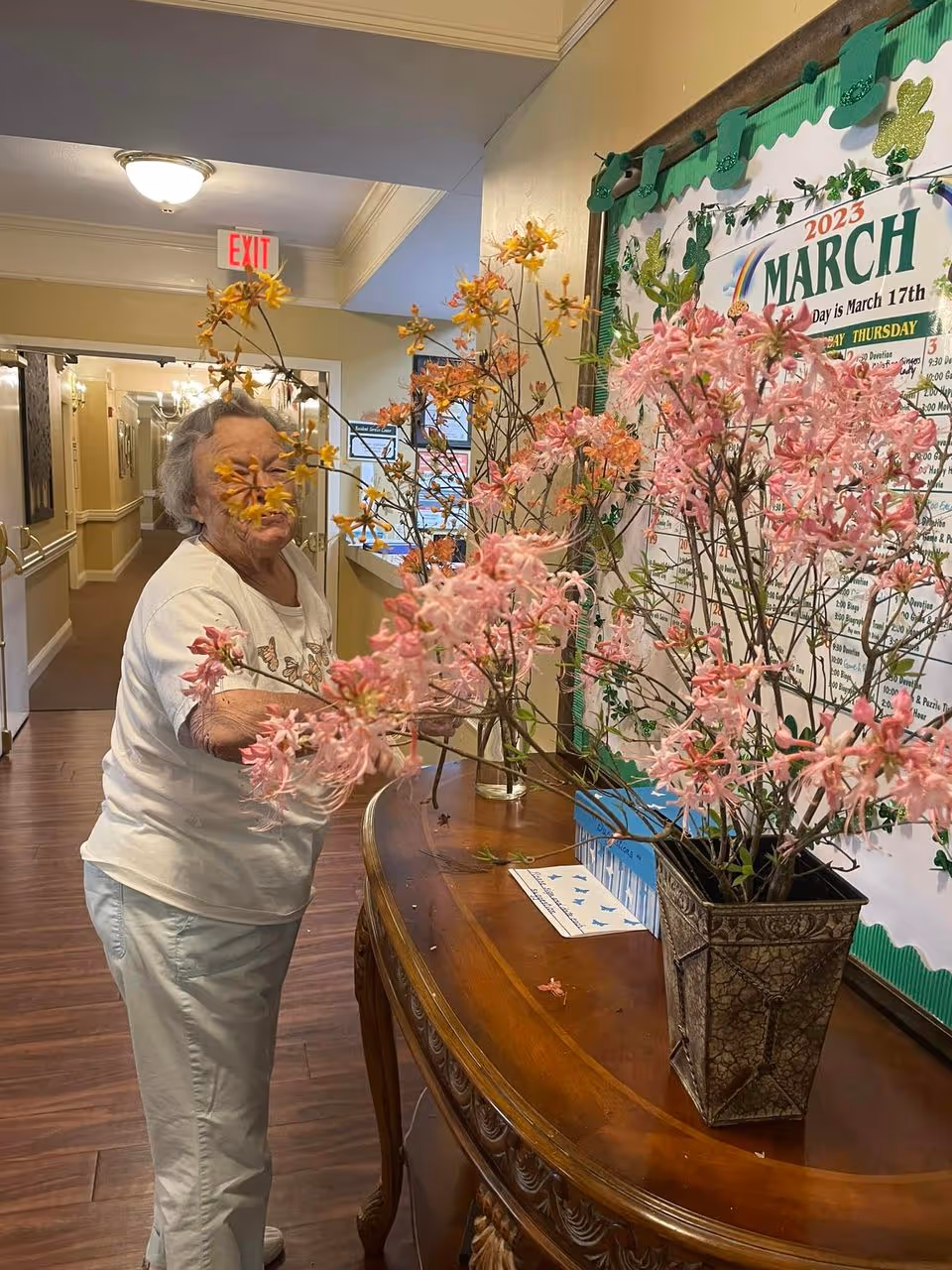 An elderly woman arranging pink and yellow flowers on a wooden table in a hallway of a senior care facility. Behind her is a bulletin board with a March 2023 calendar and green shamrock decorations. The hallway has wooden flooring, cream-colored walls, and an exit sign above a door in the background.