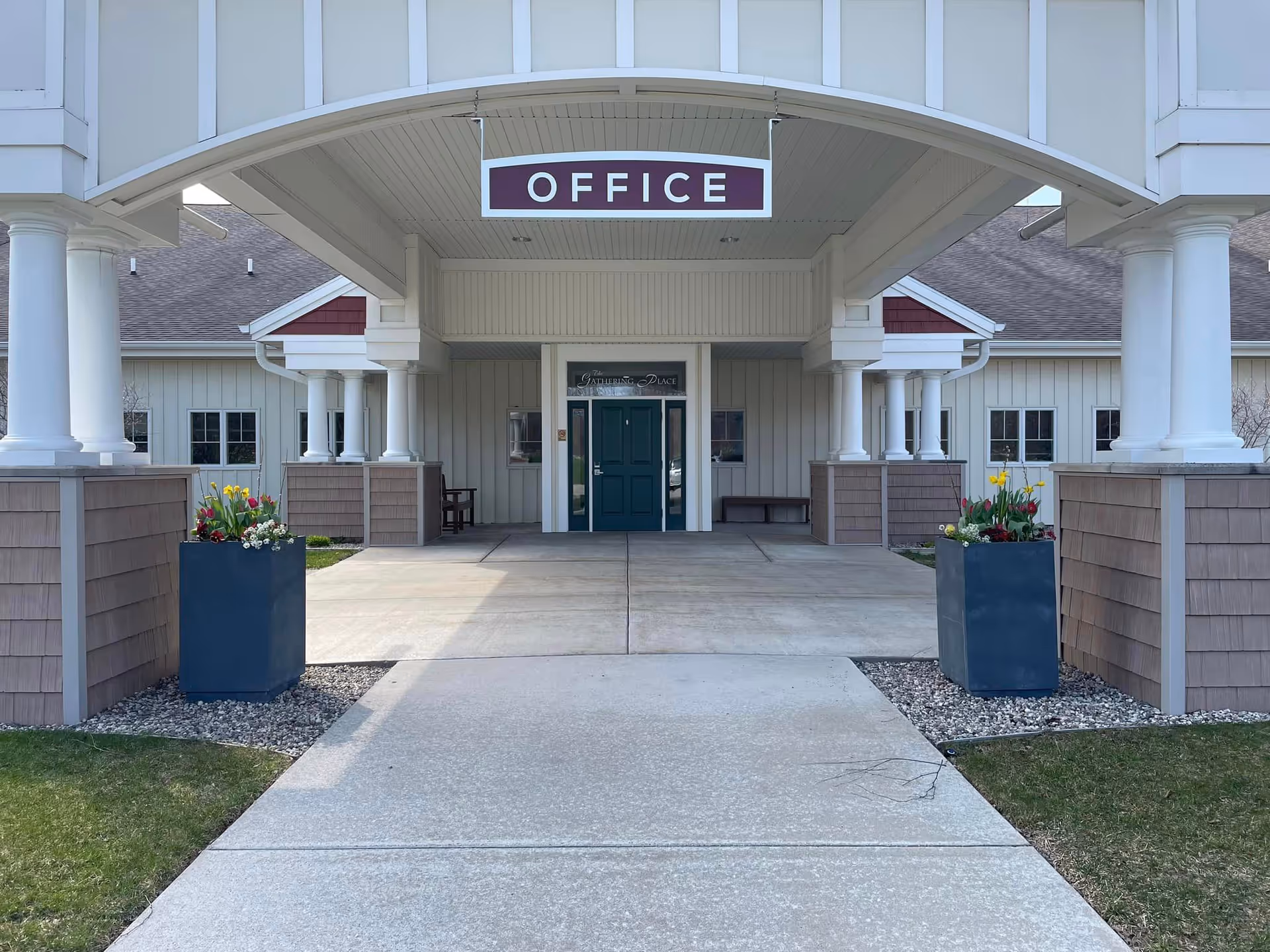 Entrance to a building with a covered walkway supported by white columns. A sign hanging from the ceiling reads 'OFFICE'. Two large planters with colorful flowers flank the entrance. The door is green and above it is a sign that says 'The Gathering Place'.