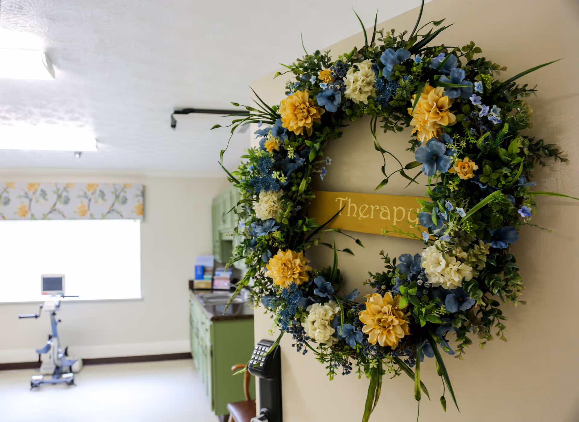 Decorative floral wreath with a 'Therapy' sign hanging on a wall in a therapy/exercise room.