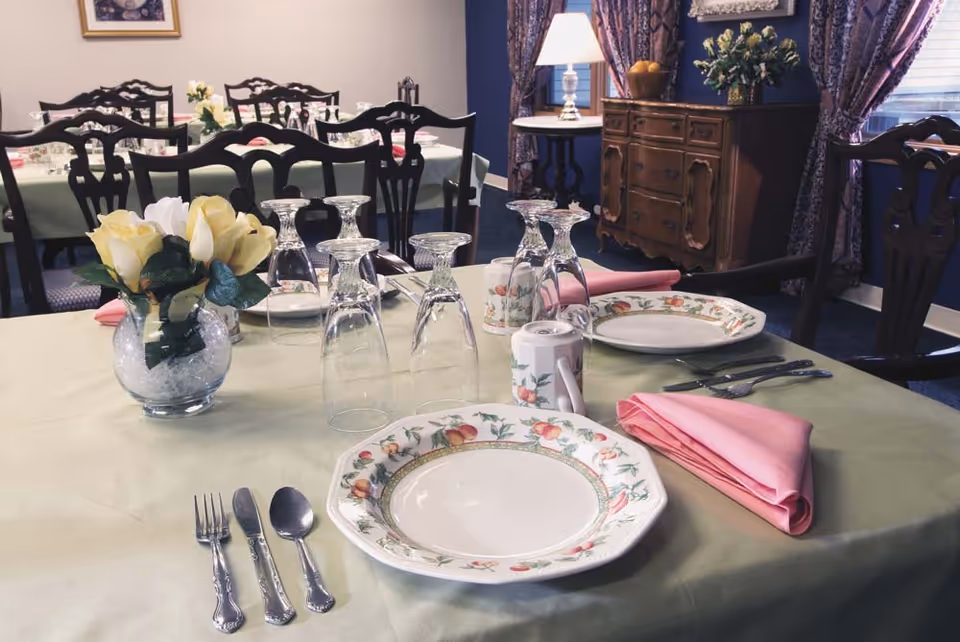 A formal dining room table set with floral china, upside-down wine glasses, pink napkins, silverware, and a small floral centerpiece.