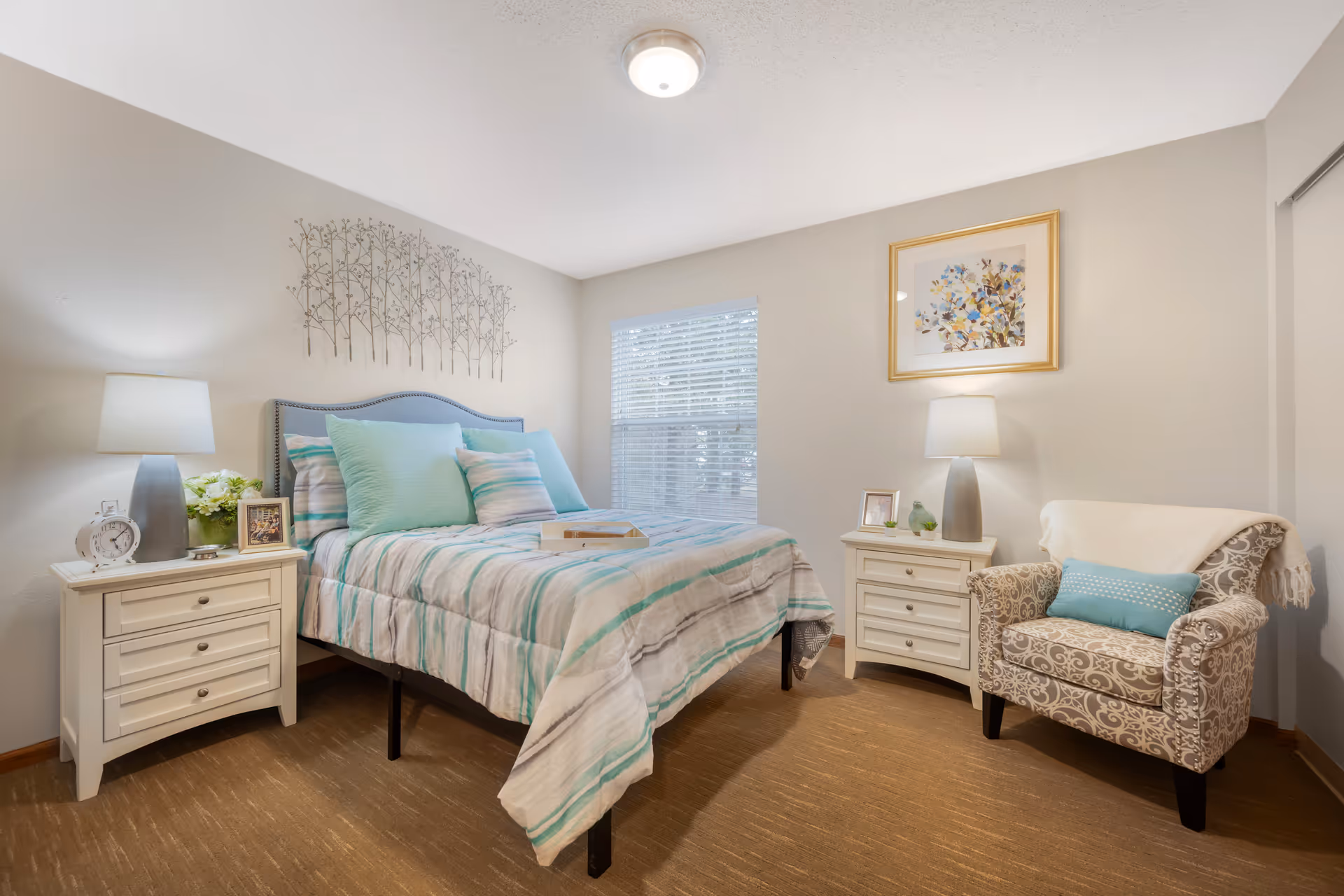 A cozy bedroom with a bed featuring a striped comforter and multiple pillows in shades of blue and white. There are two white nightstands on either side of the bed, each with a lamp, framed photos, and decorative items. A patterned armchair with a blue pillow and a white throw blanket is positioned to the right. A window with blinds lets in natural light, and the walls are decorated with a metal tree wall art above the bed and a framed floral painting above the nightstand on the right.