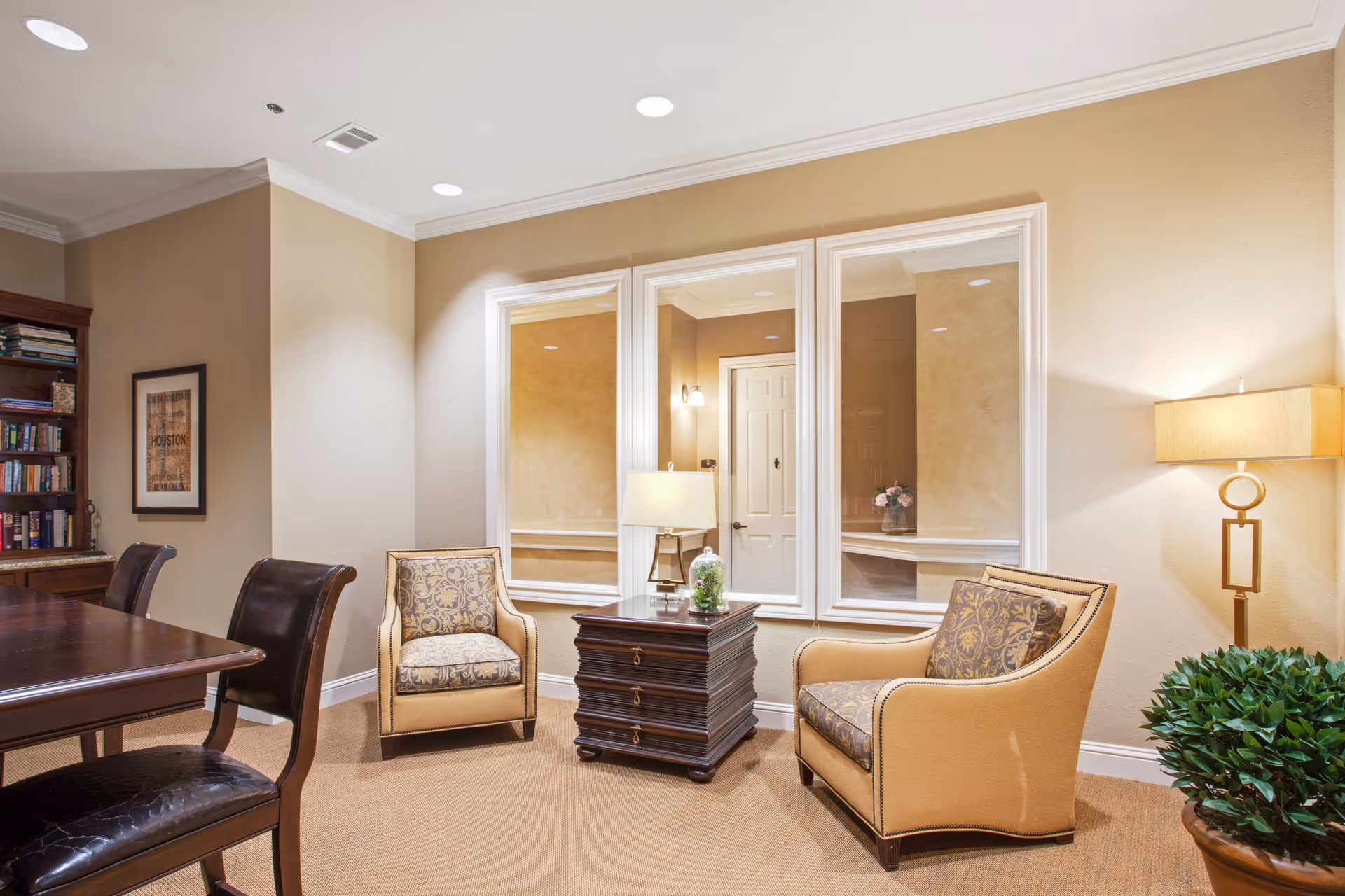 A cozy interior room with two upholstered armchairs featuring patterned cushions, a dark wooden side table with a lamp and decorative plant, a floor lamp, and a bookshelf filled with books. The walls are beige with white trim and three large windows reflect the room's lighting.