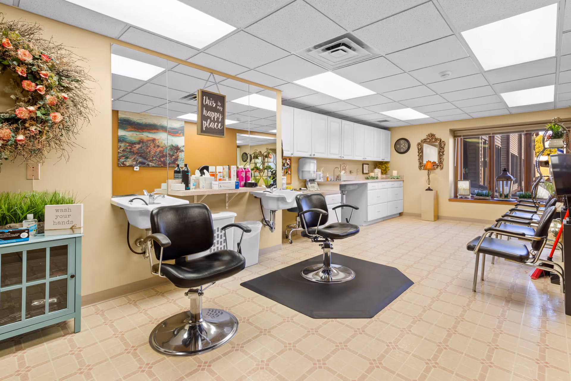 Interior of a senior living facility's hair salon with two black salon chairs in front of sinks and a large mirror. The room has beige walls, a floral wreath, a sign that says 'this is my happy place,' and a small cabinet with a 'wash your hands' sign. There are additional chairs lined up near a window with plants and decorative items on the windowsill.