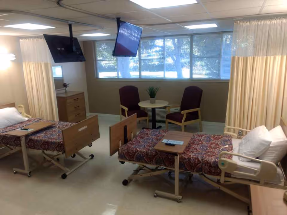 A shared patient room in a healthcare facility with two hospital beds, each with a patterned bedspread and a small overbed table. There are two chairs and a small round table near a large window with blinds. Two wall-mounted televisions are positioned above the beds. Privacy curtains are partially drawn around each bed.