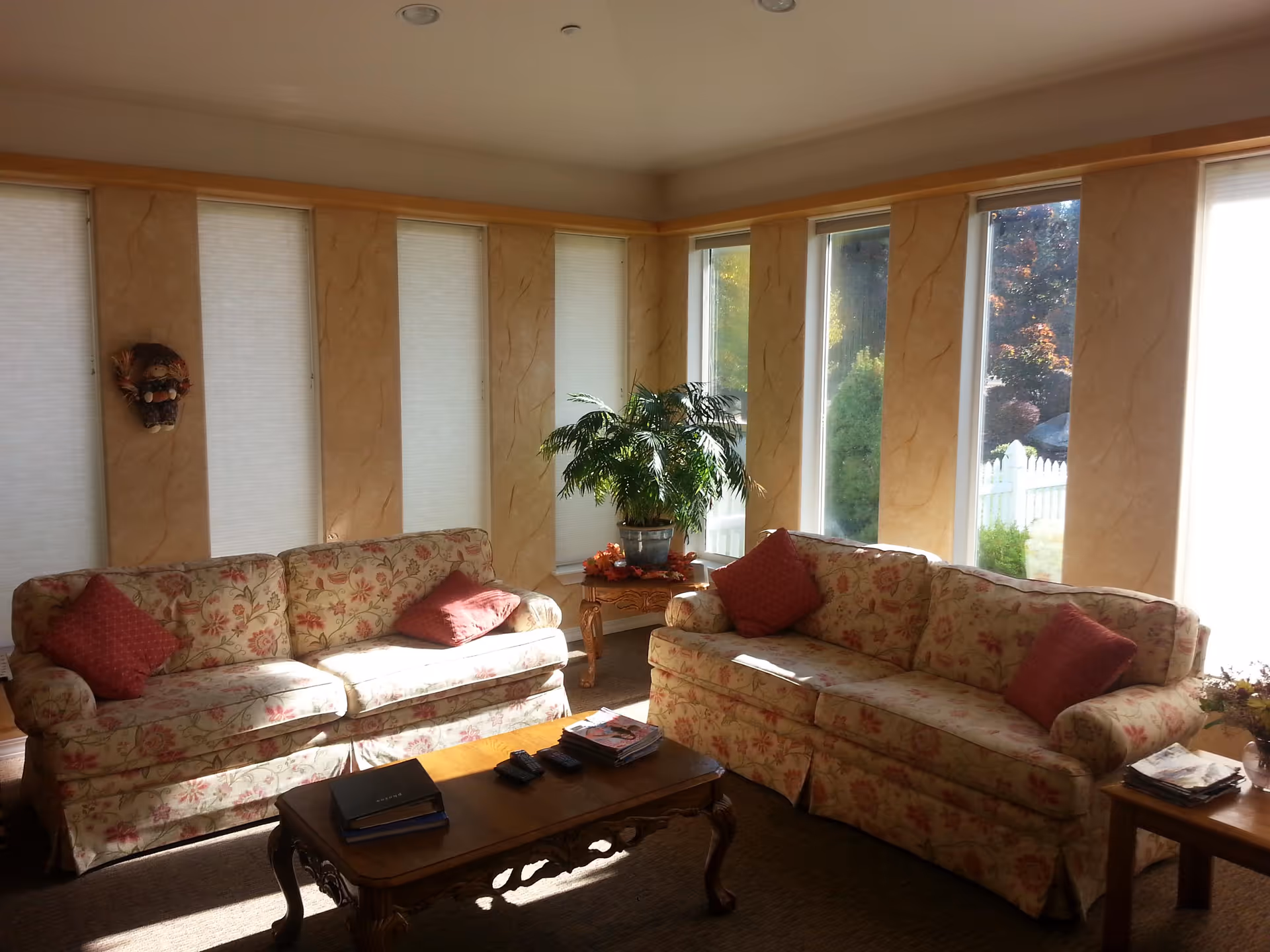 Sunlit living room with two floral-patterned sofas facing a wooden coffee table, tall narrow windows, and a potted plant between the sofas.
