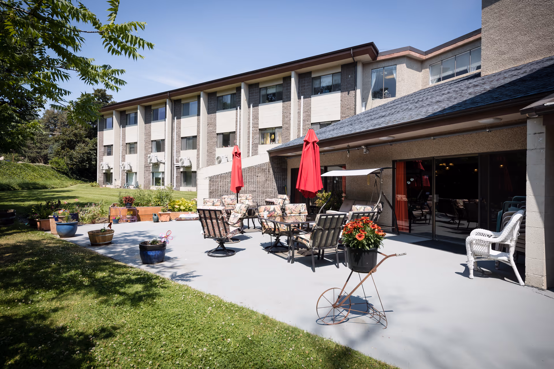 Outdoor patio area at Cherry Heights Living with several cushioned chairs and tables under red umbrellas, surrounded by potted plants and flowers. The patio is adjacent to a multi-story building with large windows, and there is a grassy lawn and trees nearby under a clear blue sky.