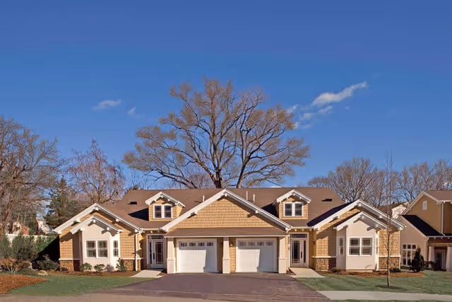 Exterior view of a residential building with two attached units, each featuring a garage door, windows, and a front entrance. The building has a brown shingled roof and beige siding with white trim. There are leafless trees in the background under a clear blue sky.