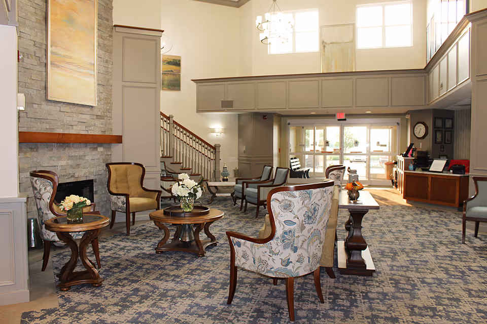 Bright senior living lobby with upholstered chairs and tables arranged around a fireplace, a reception desk, and a staircase under high windows.