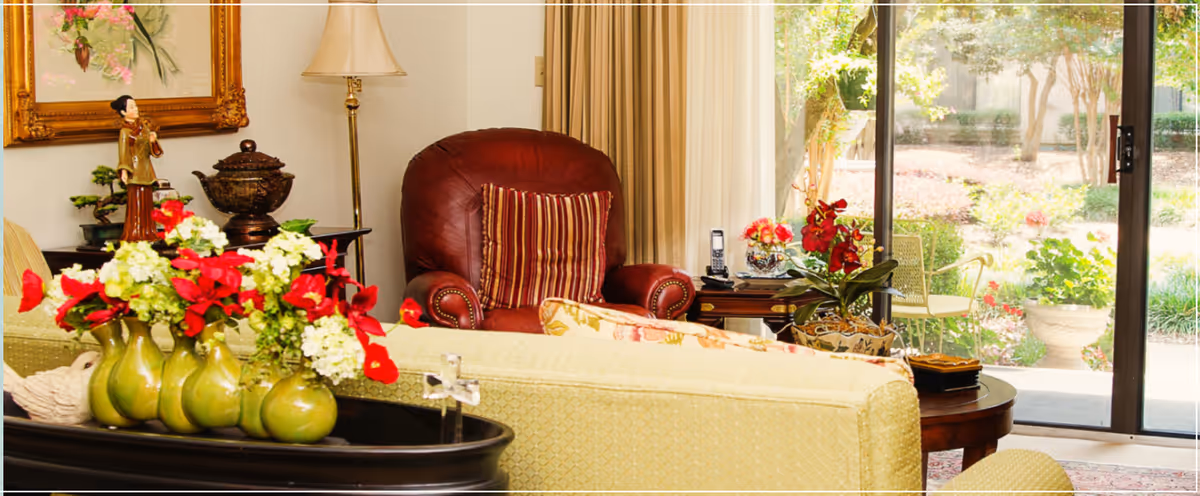 A cozy living room with a beige sofa adorned with patterned cushions, a red leather armchair with a striped pillow, a side table with a lamp, and a large window or sliding glass door showing a garden outside. There are decorative flower arrangements on a black tray and on the side table.