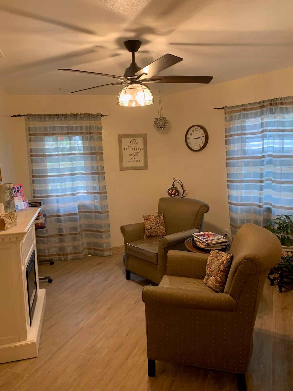 A cozy living room with two upholstered armchairs with floral cushions, a small round wooden table with magazines, a wall clock, a framed sign that reads 'Do all things with Love', two windows with sheer striped curtains, a ceiling fan with a light fixture, and a white electric fireplace.