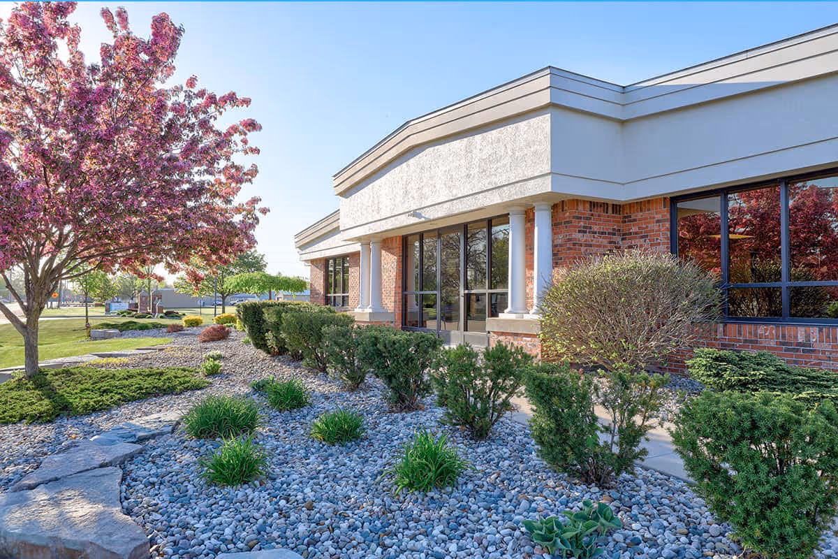 Front exterior of a brick nursing and rehab building with glass doors, landscaped rock beds, shrubs, and a blooming pink tree.