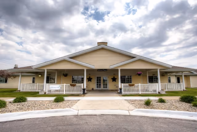 Front exterior view of a single-story senior living facility building with a covered entrance, white railings, decorative stars on the wall, and a sign that reads 'Pathfinder Senior Living'. The sky is partly cloudy.