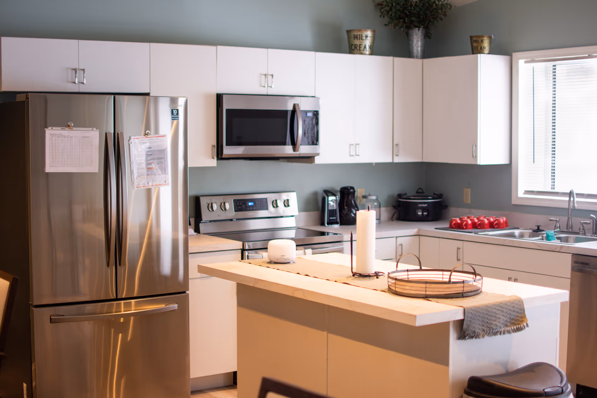 Modern kitchen with stainless steel refrigerator, microwave, and stove. White cabinets line the walls with a countertop island in the center featuring a candle and a decorative tray. A window above the sink lets in natural light, and various kitchen appliances and red mugs are visible on the counters.