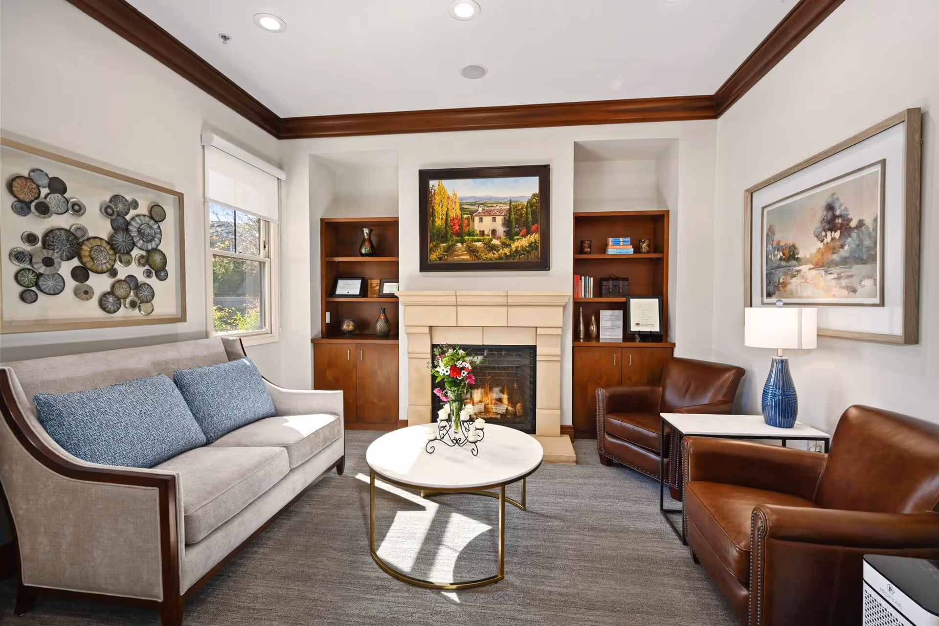 A cozy living room with a beige sofa with blue cushions on the left, two brown leather armchairs on the right, a round white coffee table with a flower vase in the center, and a fireplace with a painting above it in the background. There are built-in wooden shelves on either side of the fireplace, a window with a white roller shade on the left wall, and framed artwork on the walls. A table lamp with a blue base is on a side table next to one of the armchairs.