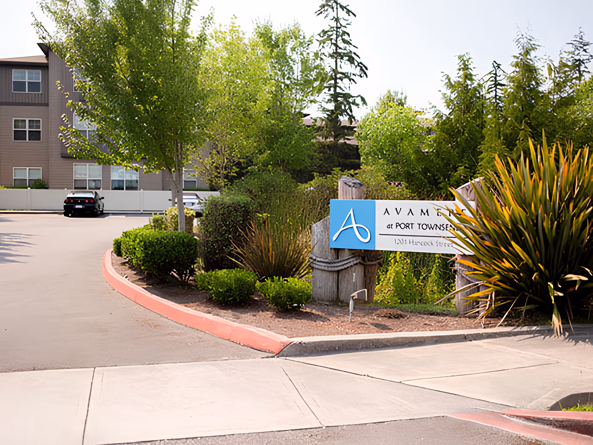 Entrance area of a senior living facility with a sign that reads 'Avamere of Port Townsend' surrounded by green bushes and trees, with a building and parked cars visible in the background.