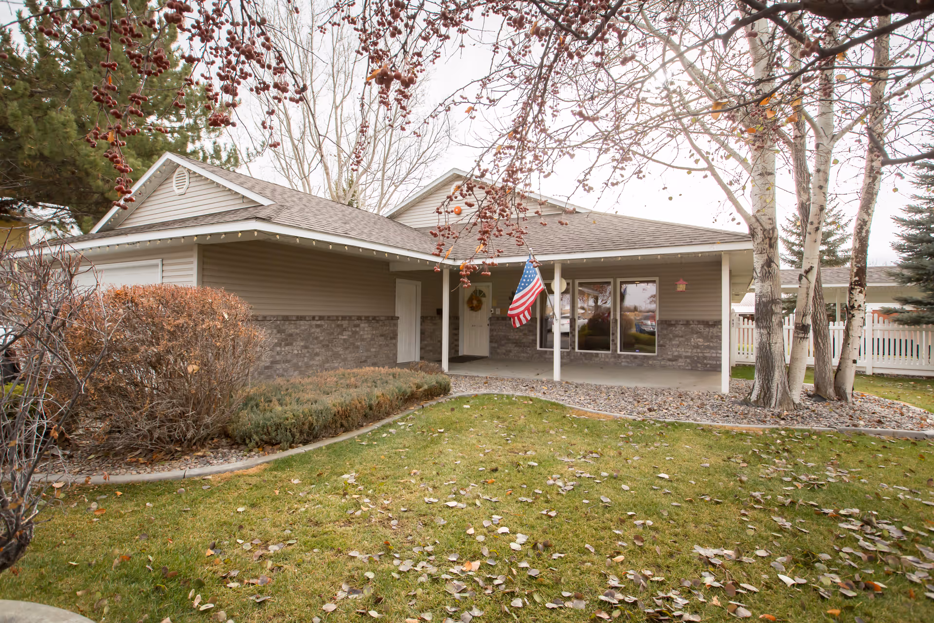 Exterior view of a single-story assisted living facility building with a covered porch, an American flag hanging near the entrance, surrounded by trees and bushes with fallen leaves on the grass.