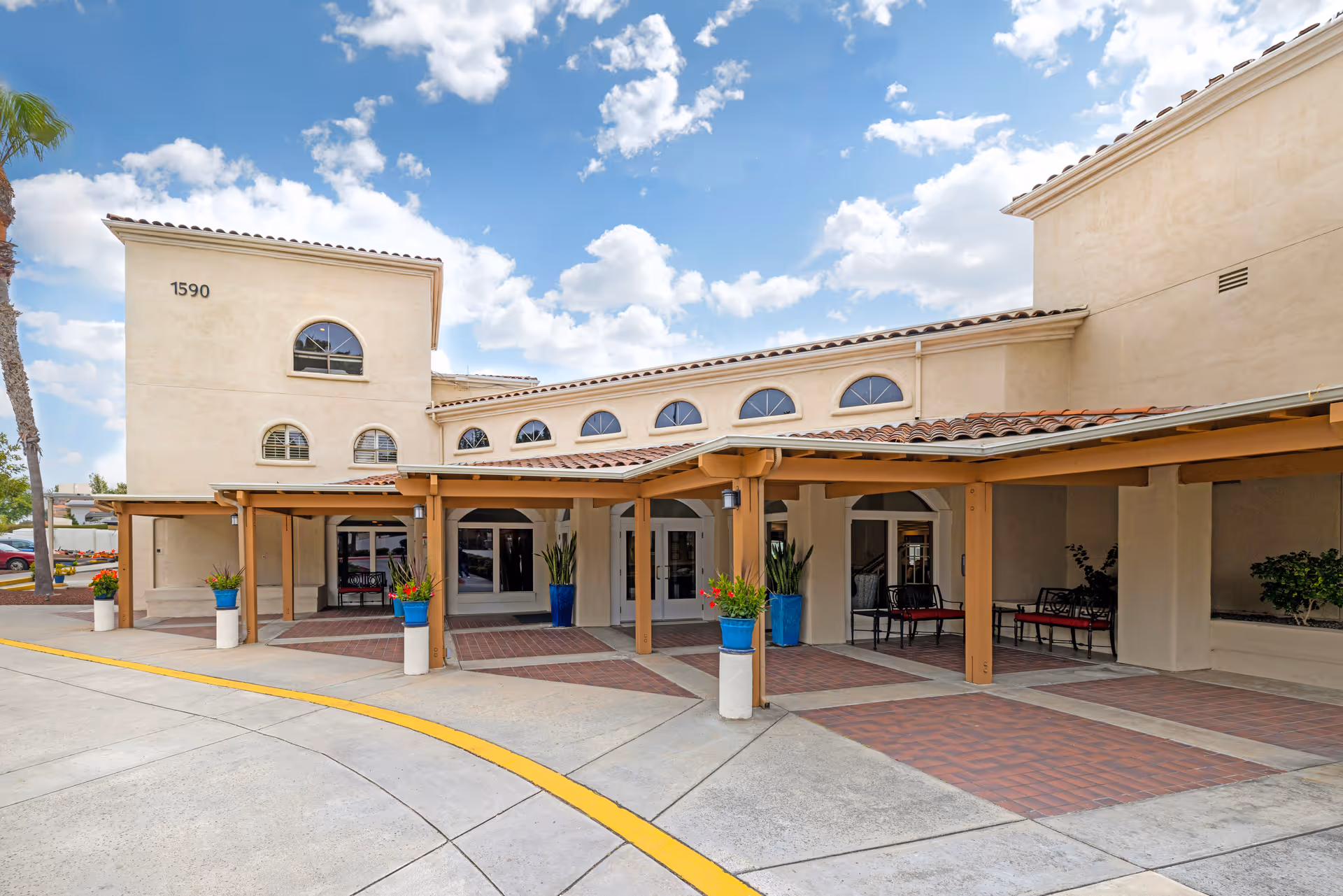 Exterior view of the entrance to a senior living facility with beige stucco walls, arched windows, a covered walkway supported by wooden beams, potted plants in blue and white containers, and a clear blue sky with some clouds.