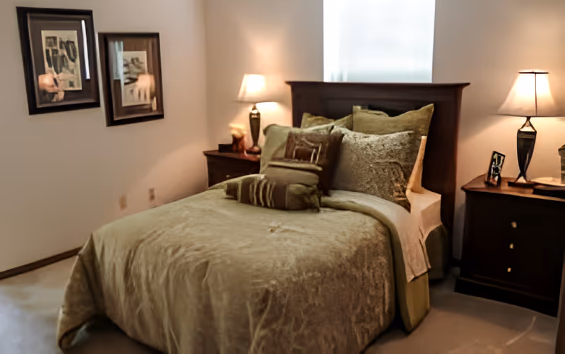 A neatly made bed with green bedding, a wooden headboard, two nightstands and lamps in a bedroom with framed artwork on the wall.