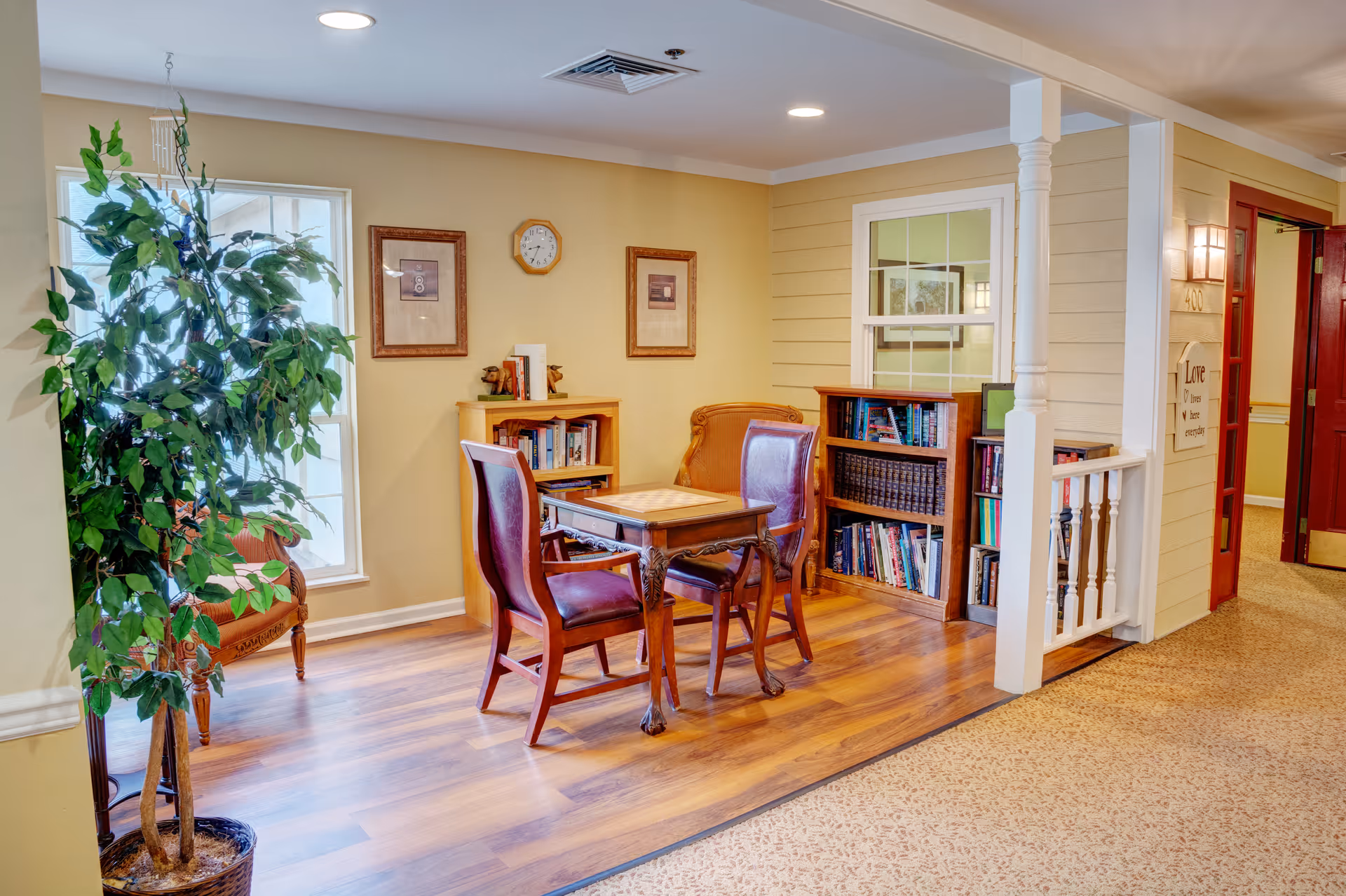 A cozy seating area in a senior living facility with two wooden chairs and a small table on a wooden floor. There are bookshelves filled with books against the walls, framed pictures and a clock hanging on the wall, a large potted plant near a window, and a hallway with red doors and a wall-mounted light fixture.