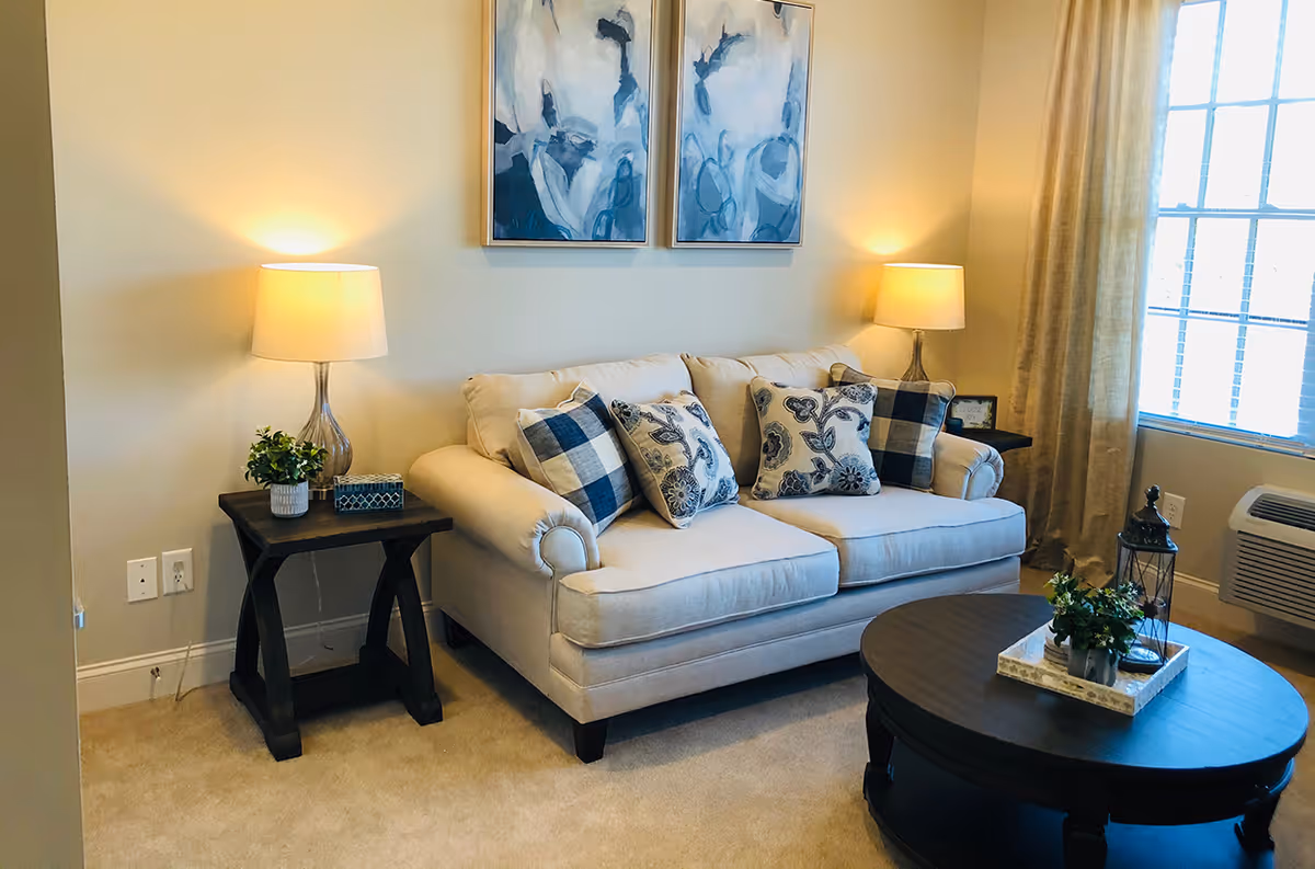 A cozy living room with a beige sofa adorned with patterned cushions, two side tables each holding a lamp and decorative items, a round dark wooden coffee table with a tray and plants, two abstract blue and white paintings on the wall, and a window with beige curtains letting in natural light.