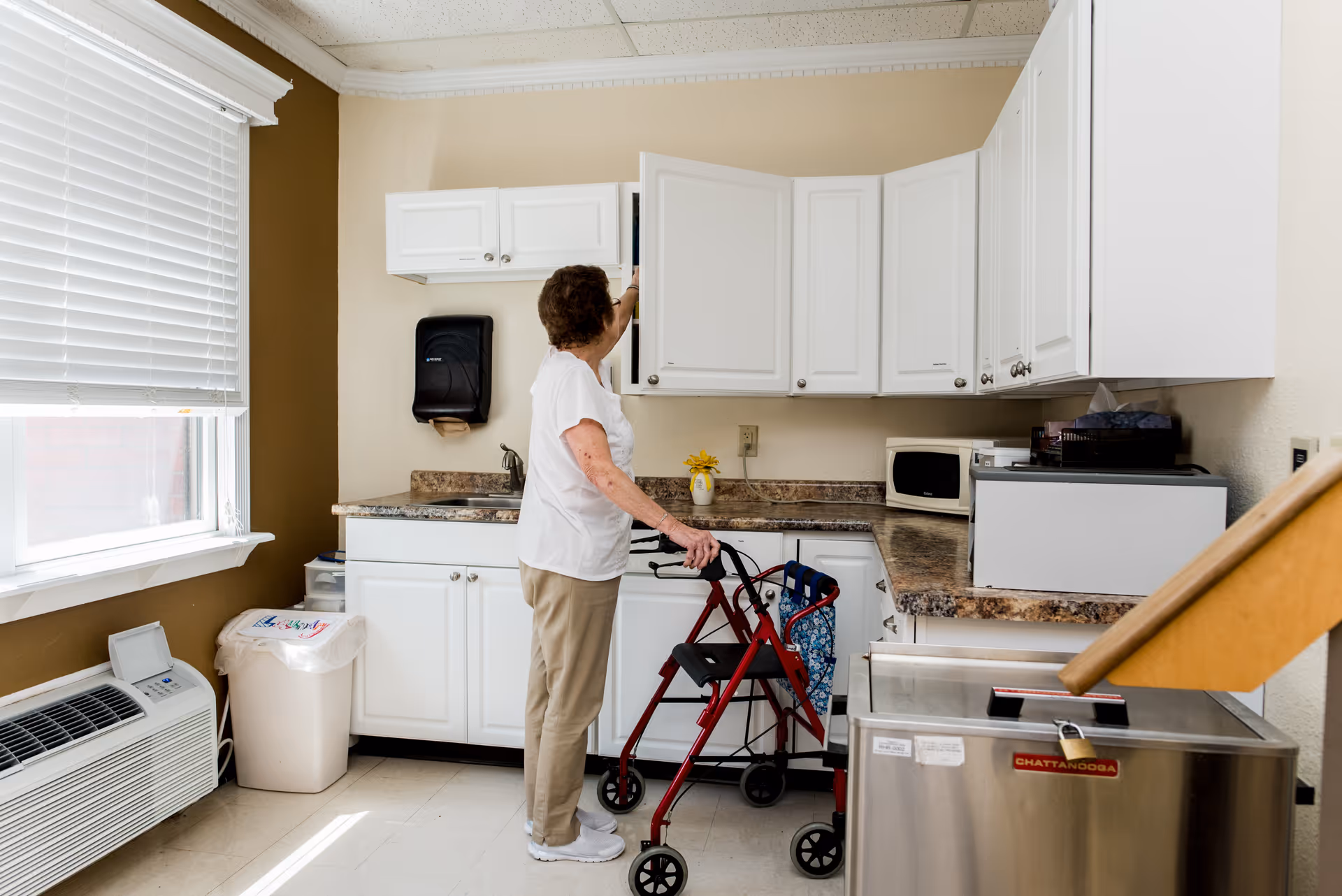 An elderly woman using a red walker reaches into a white kitchen cabinet in a small kitchenette area with white cabinets, a microwave, and a countertop. There is a window with blinds, a trash can, and an air conditioning unit visible in the room.